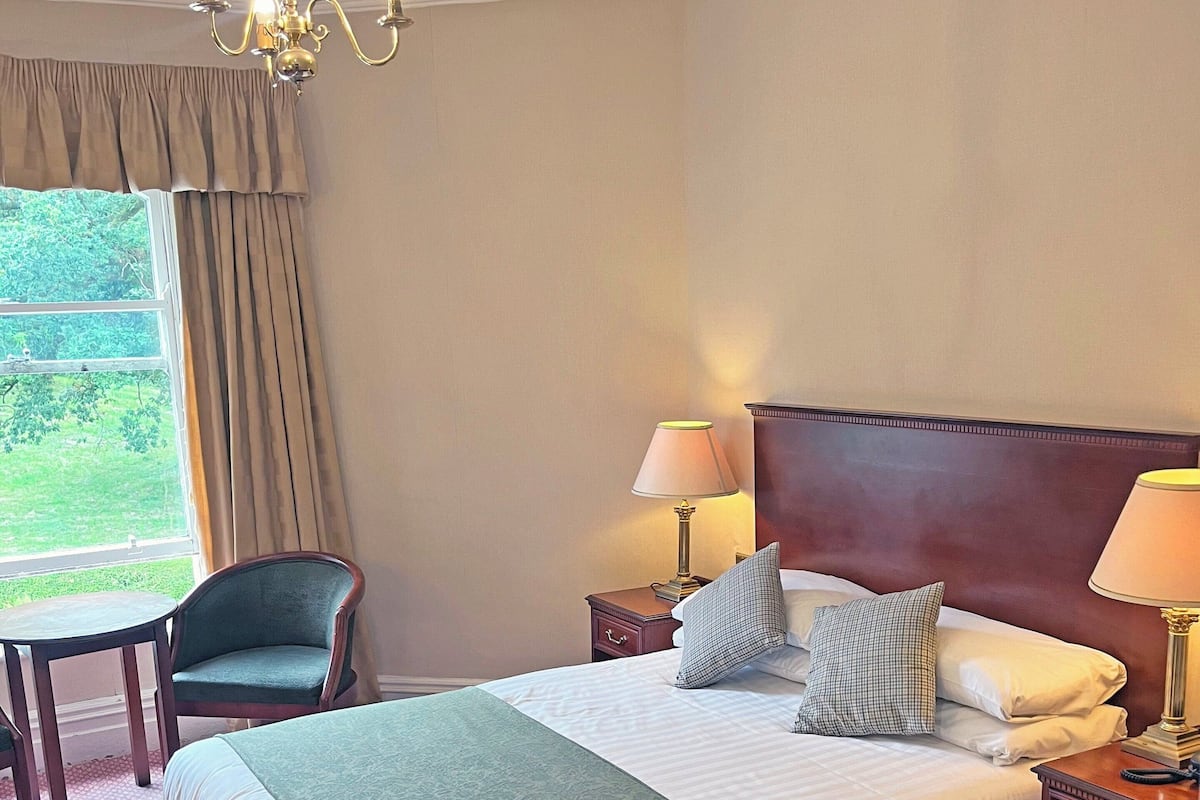 A hotel room with a large bed, two bedside tables, a chair, a chandelier, and a window with curtains.