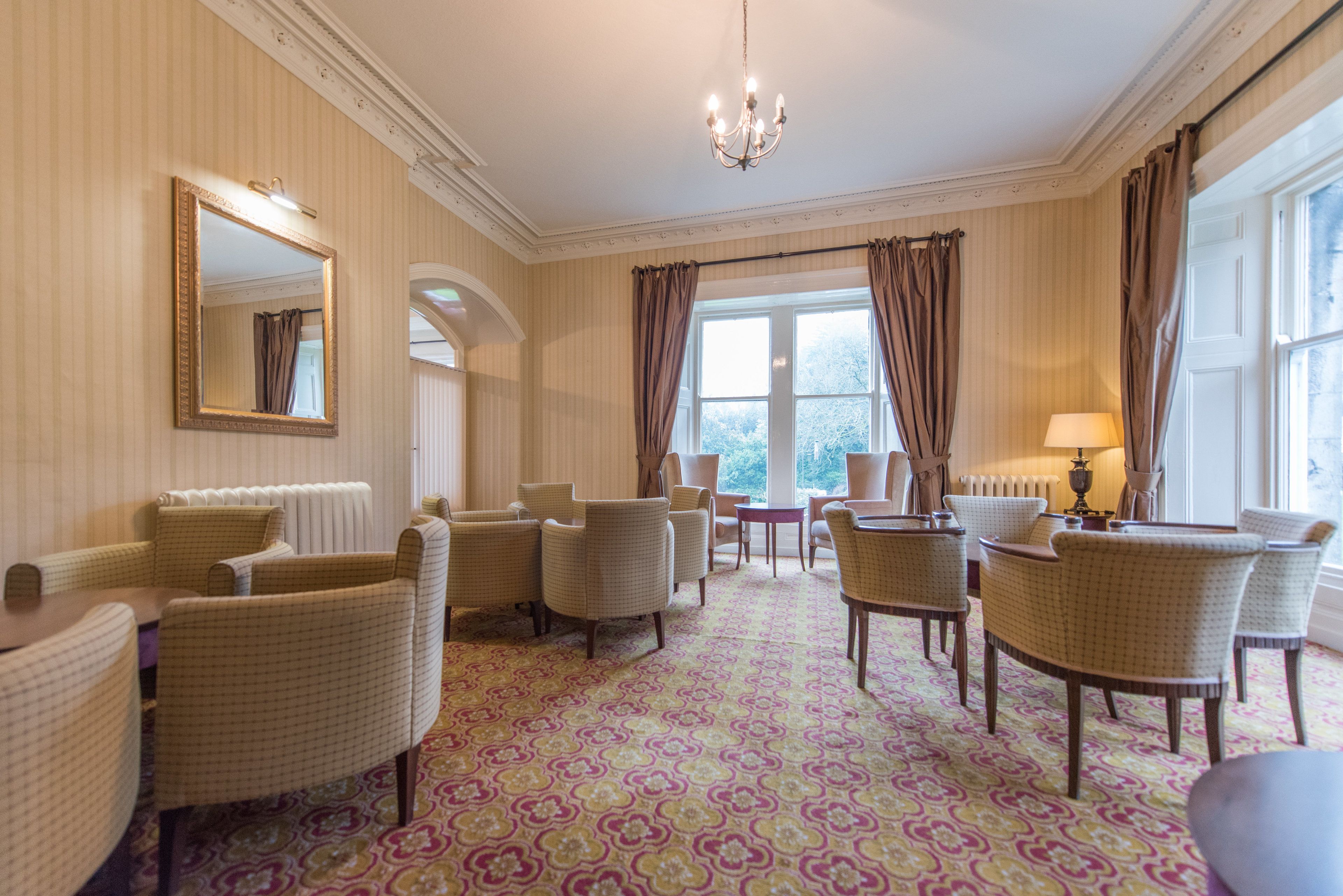 Hotelkamer met een kamerbreed tapijt, meerdere rieten stoelen, een kroonluchter en een groot raam met gordijnen.