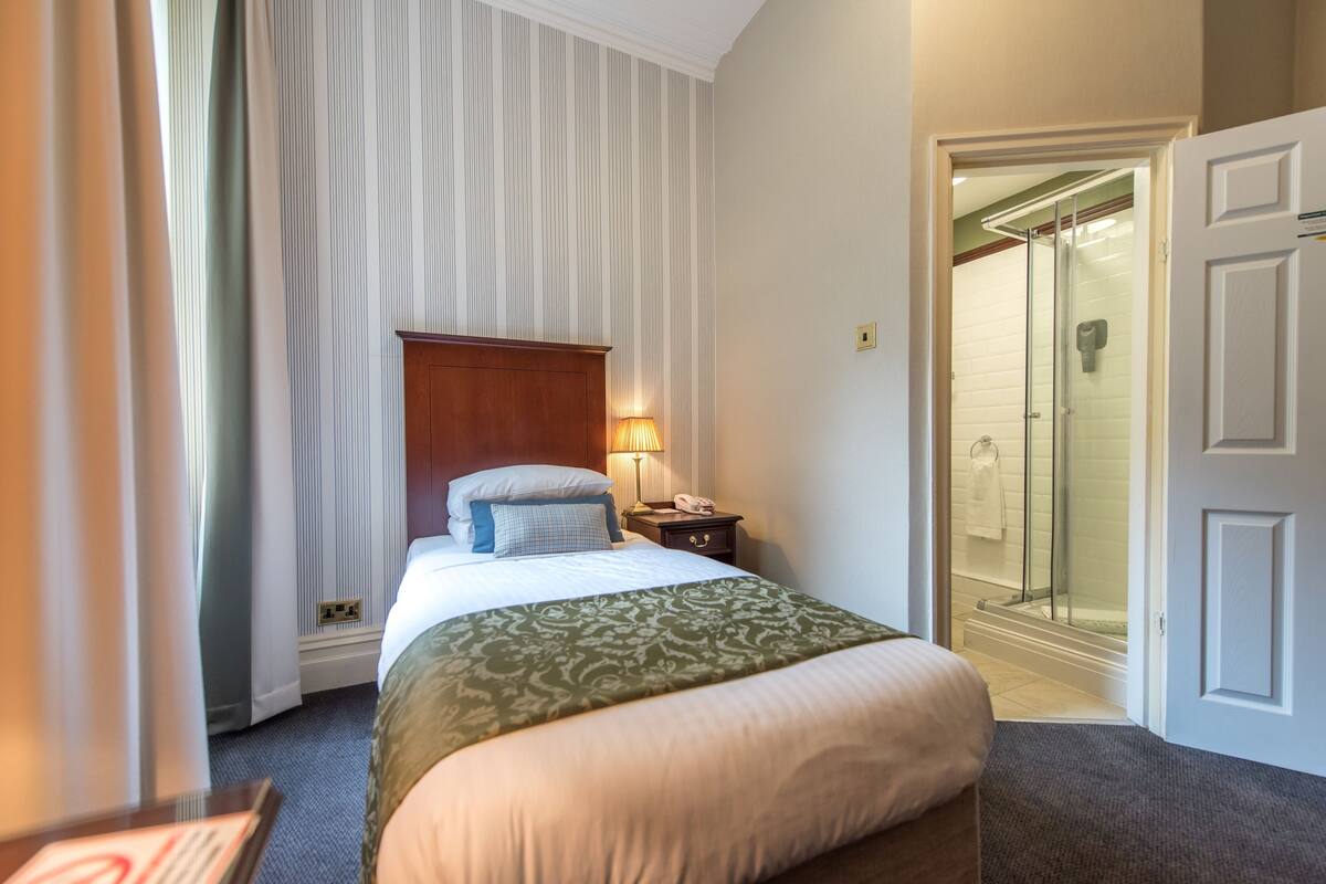 A hotel room with a bed, a nightstand, a lamp, a shower area, and a door.