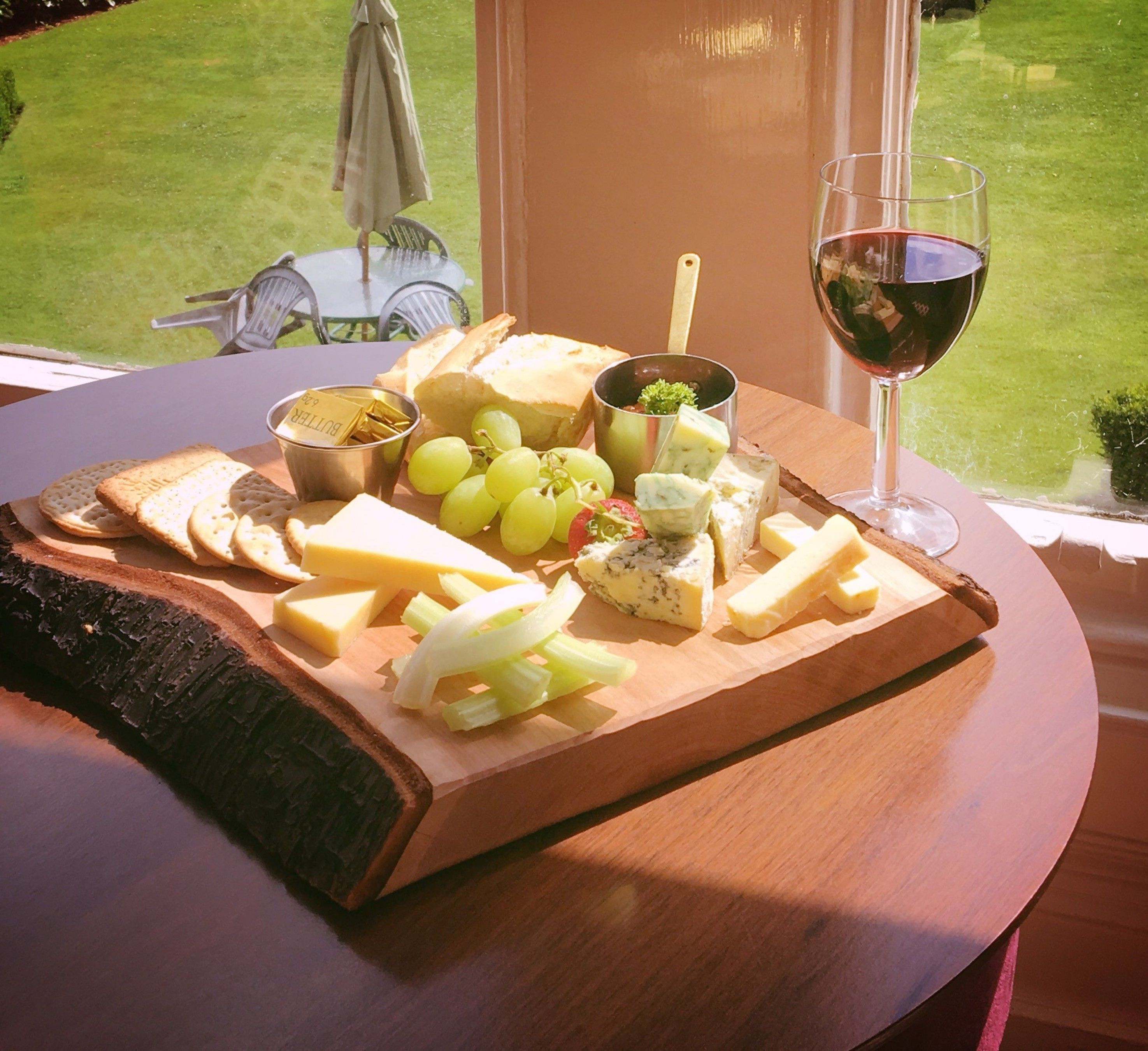 A wooden board with cheese, crackers, grapes, and a glass of wine on a table by a window.