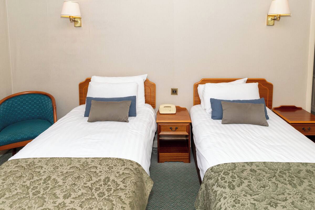 A hotel room with two single beds, a wooden nightstand, a telephone, and a teal chair.