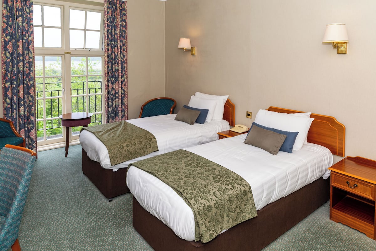 A hotel room with two beds, a small table, a chair, and a view of greenery through the window.
