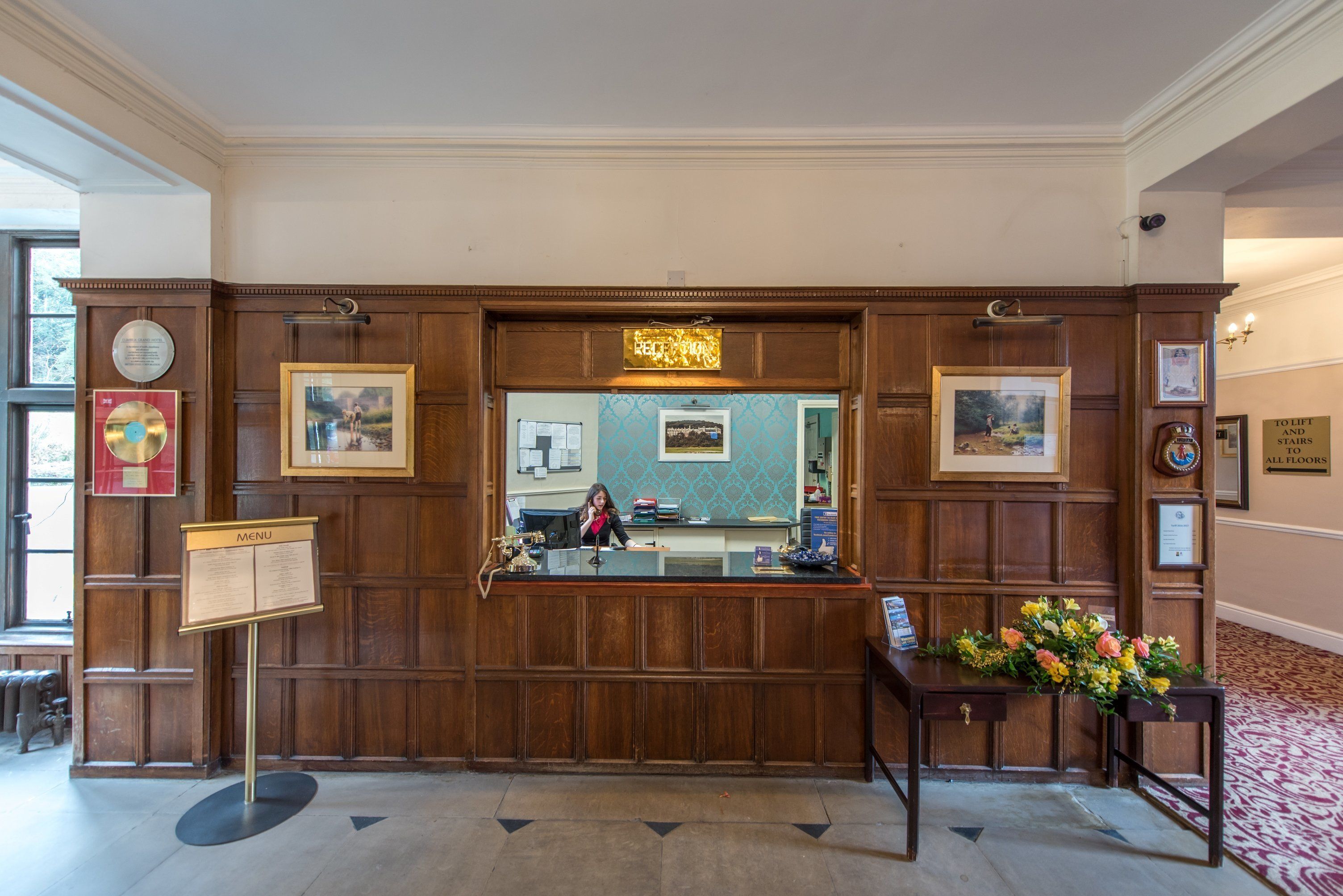 A reception area with a wooden reception desk, framed pictures, a menu stand, and a floral arrangement.