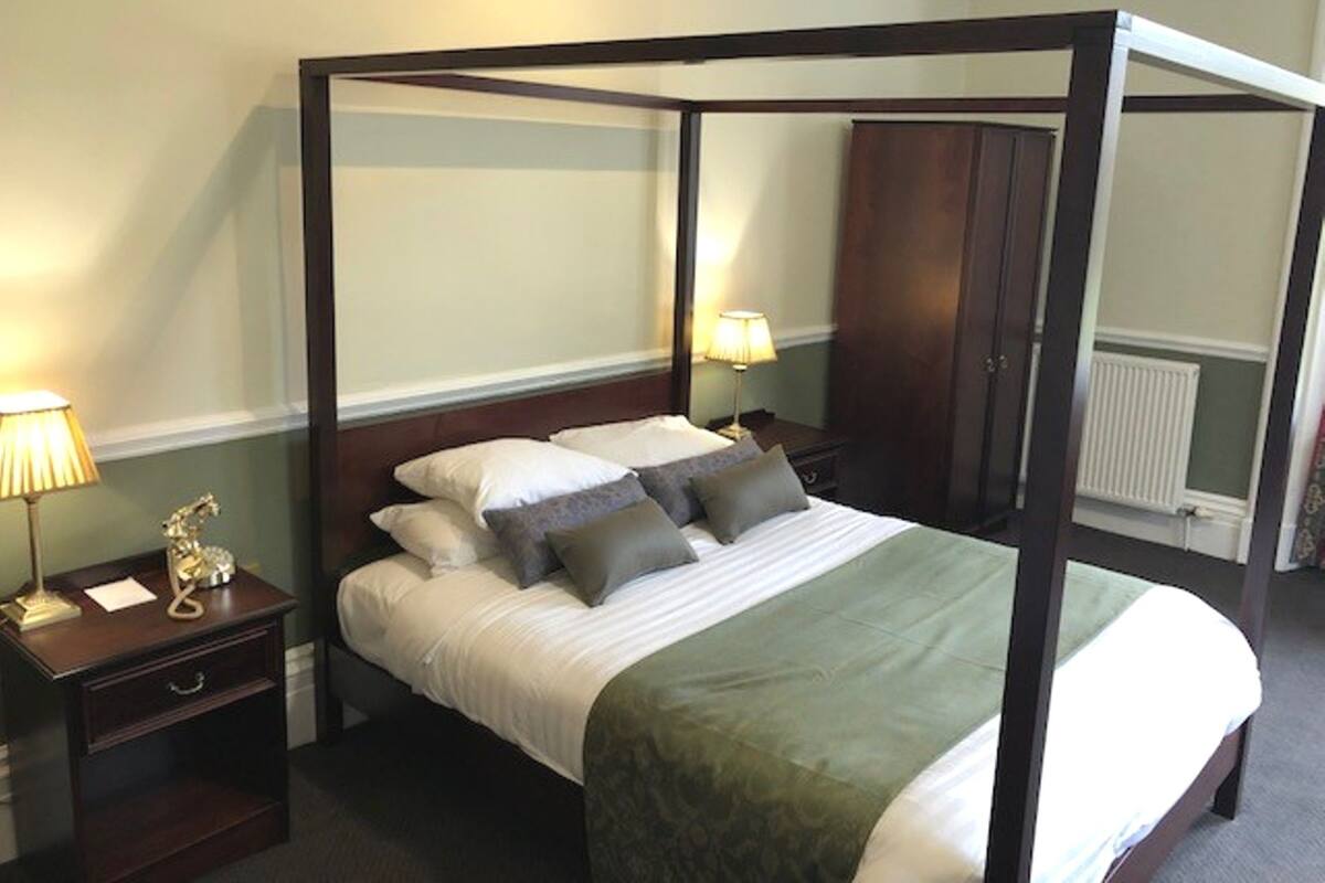 A four-poster bed with white bedding and multiple pillows, flanked by two bedside tables with lamps and a small decorative item.