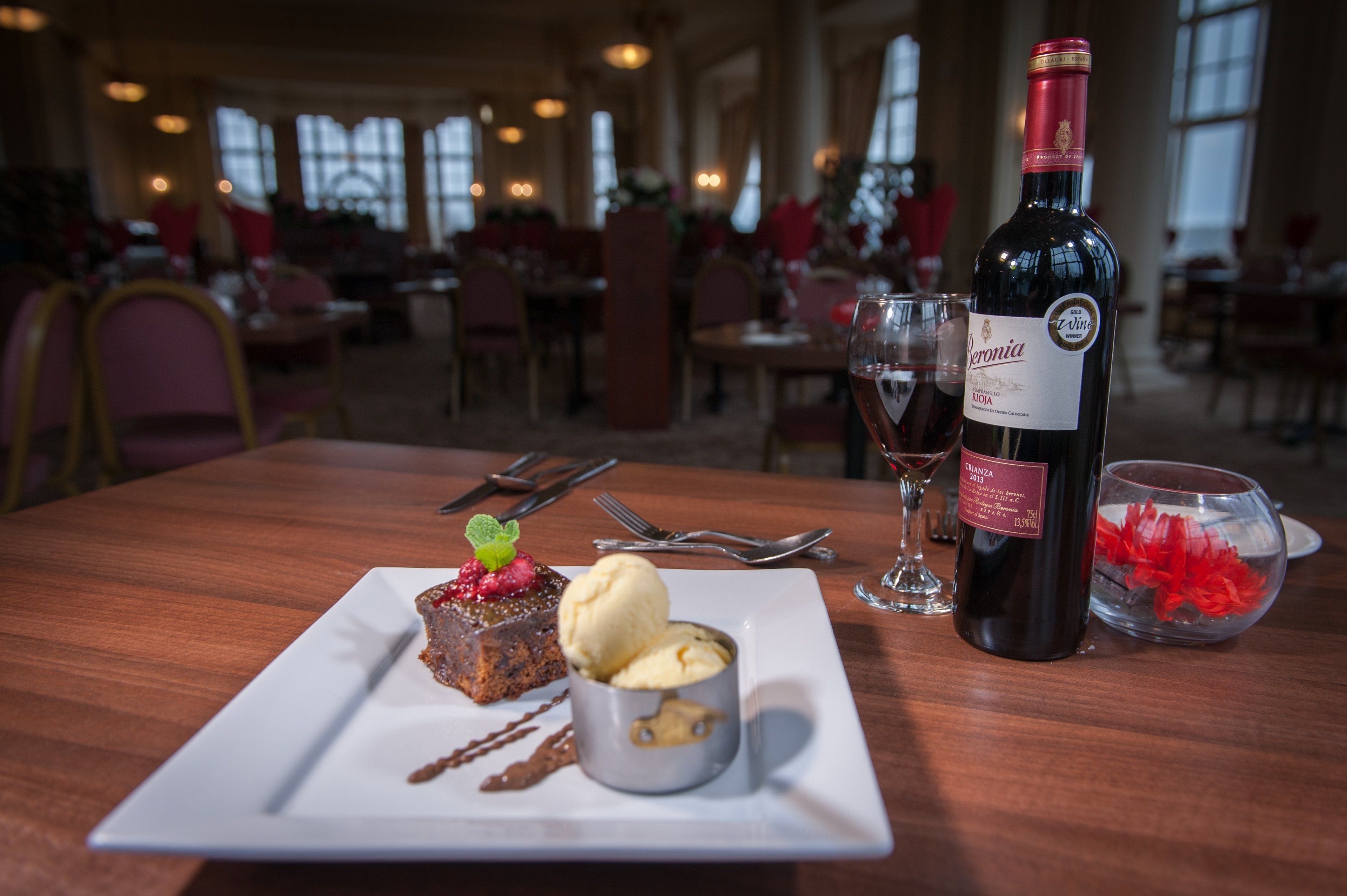 A dessert plate with a slice of cake, a scoop of ice cream, and a mint garnish on a white plate. A bottle of wine and a glass are also on the table.