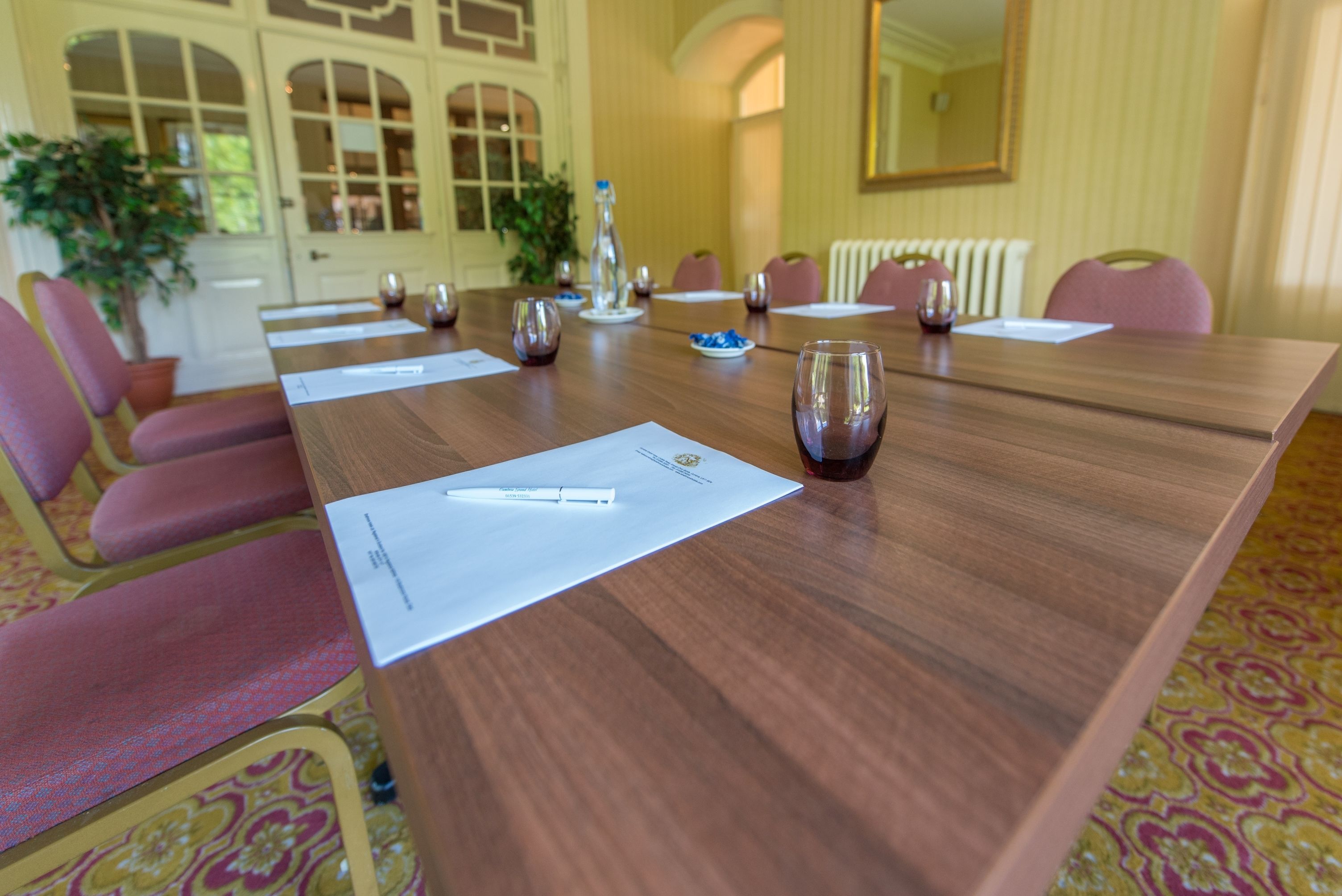 A conference room with a long wooden table, pink chairs, and a large mirror on the wall.