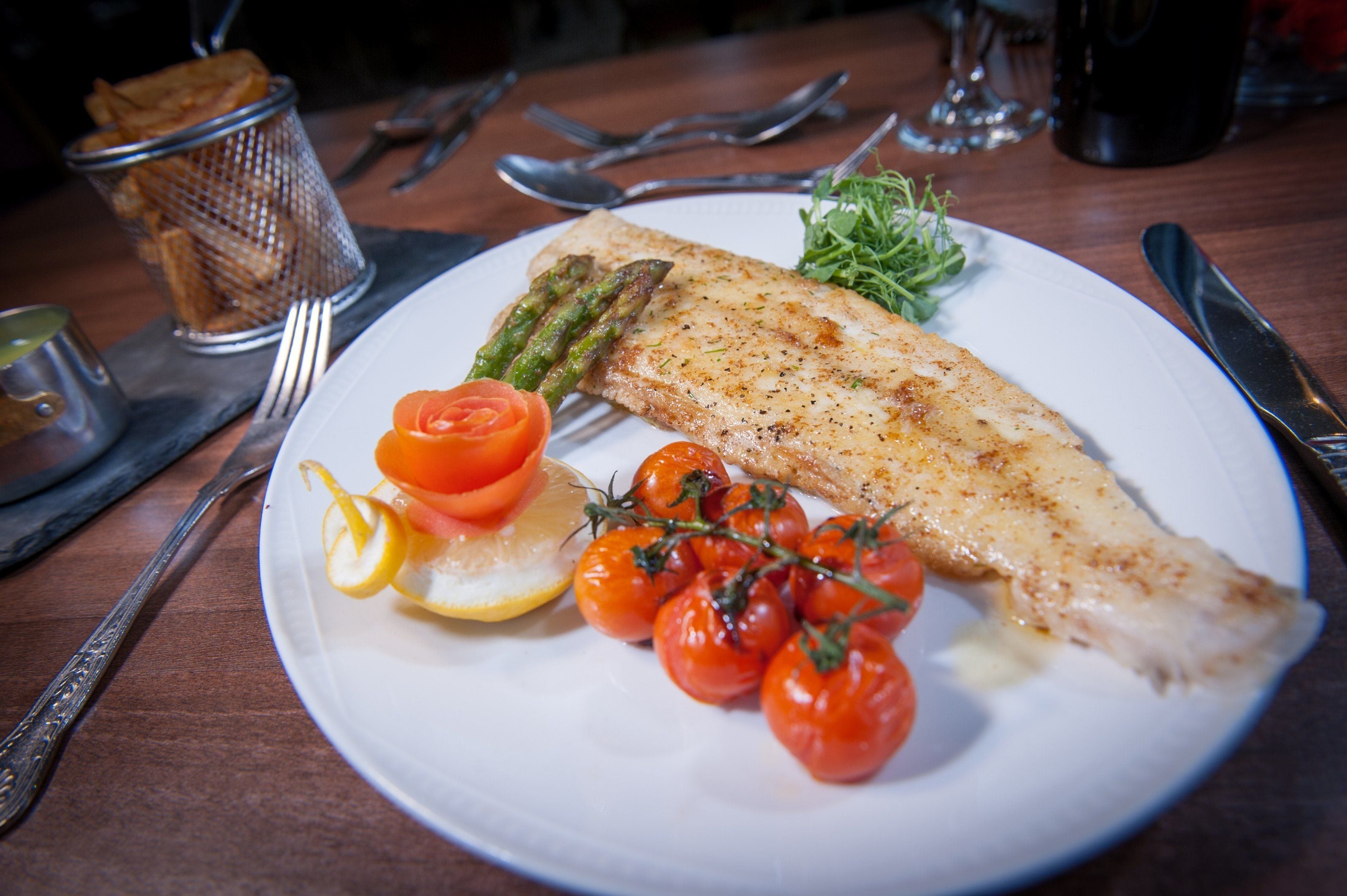A plate with a fish, asparagus, cherry tomatoes, and lemon on a wooden table.