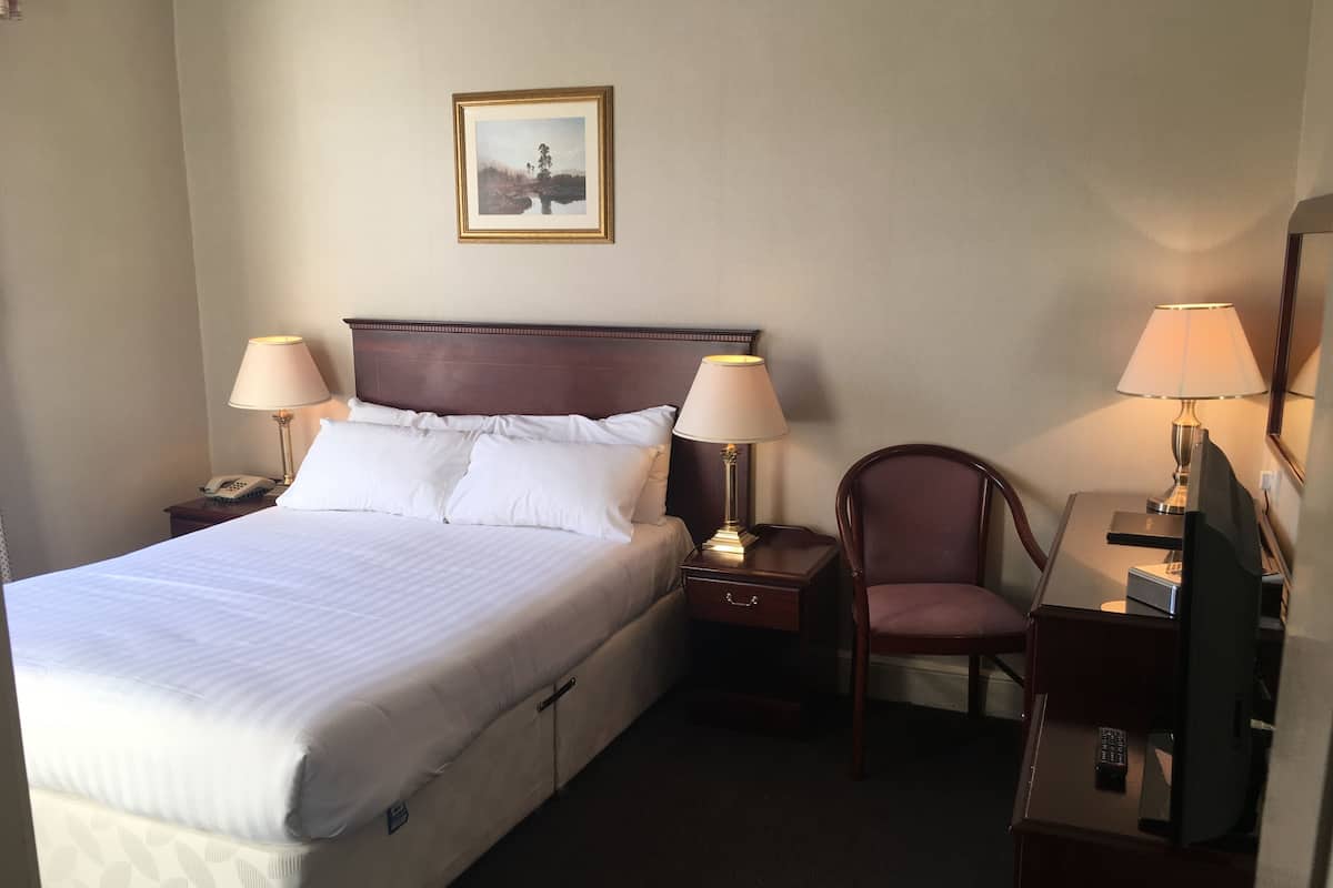 A hotel room with a large bed, two bedside tables with lamps, a chair, a desk with a computer, and a framed picture on the wall.