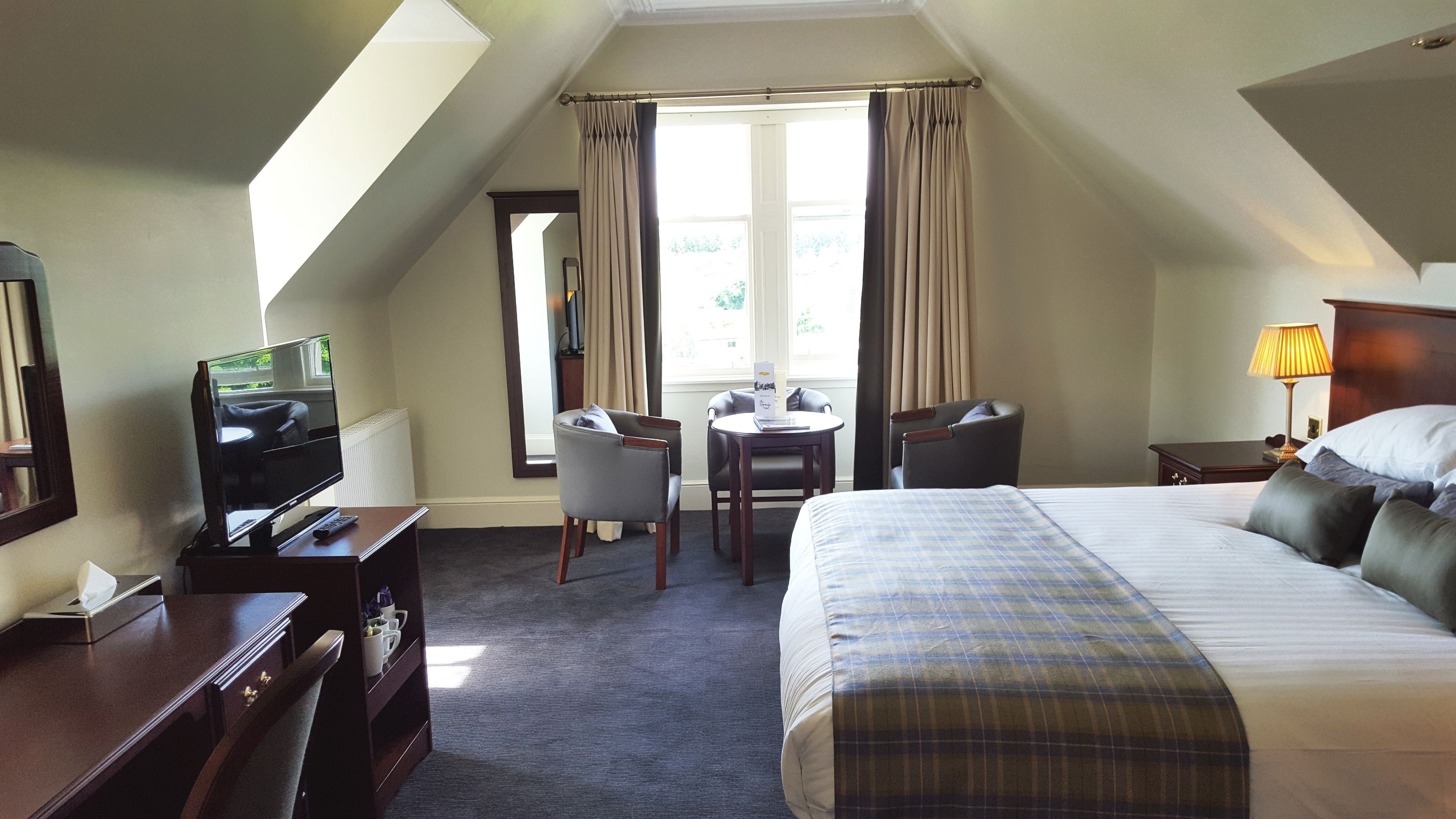 A hotel room with a bed, a desk with a chair, a television, and a window with curtains.