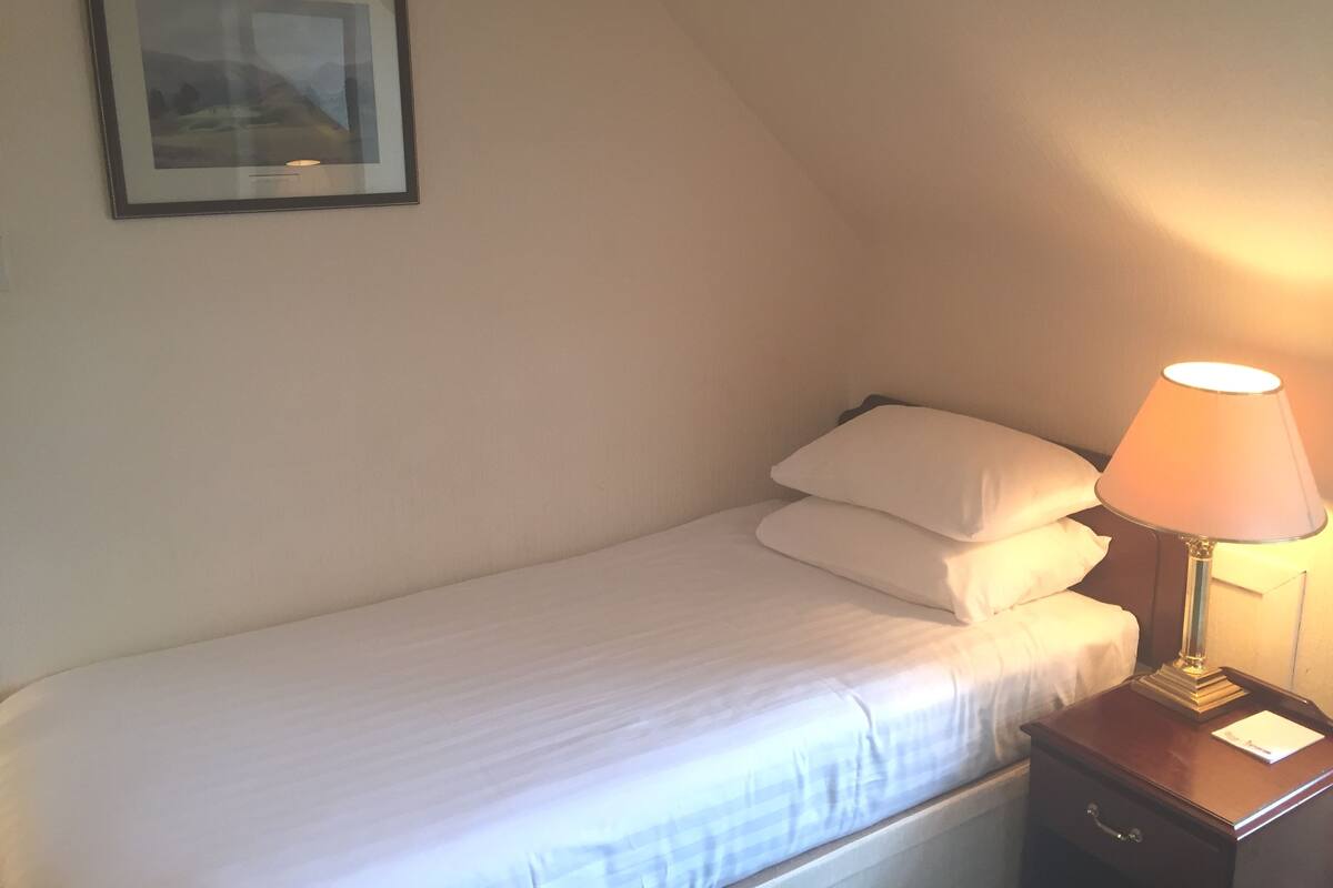A small, neatly made bed with white linens, a bedside lamp, and a framed picture on the wall.
