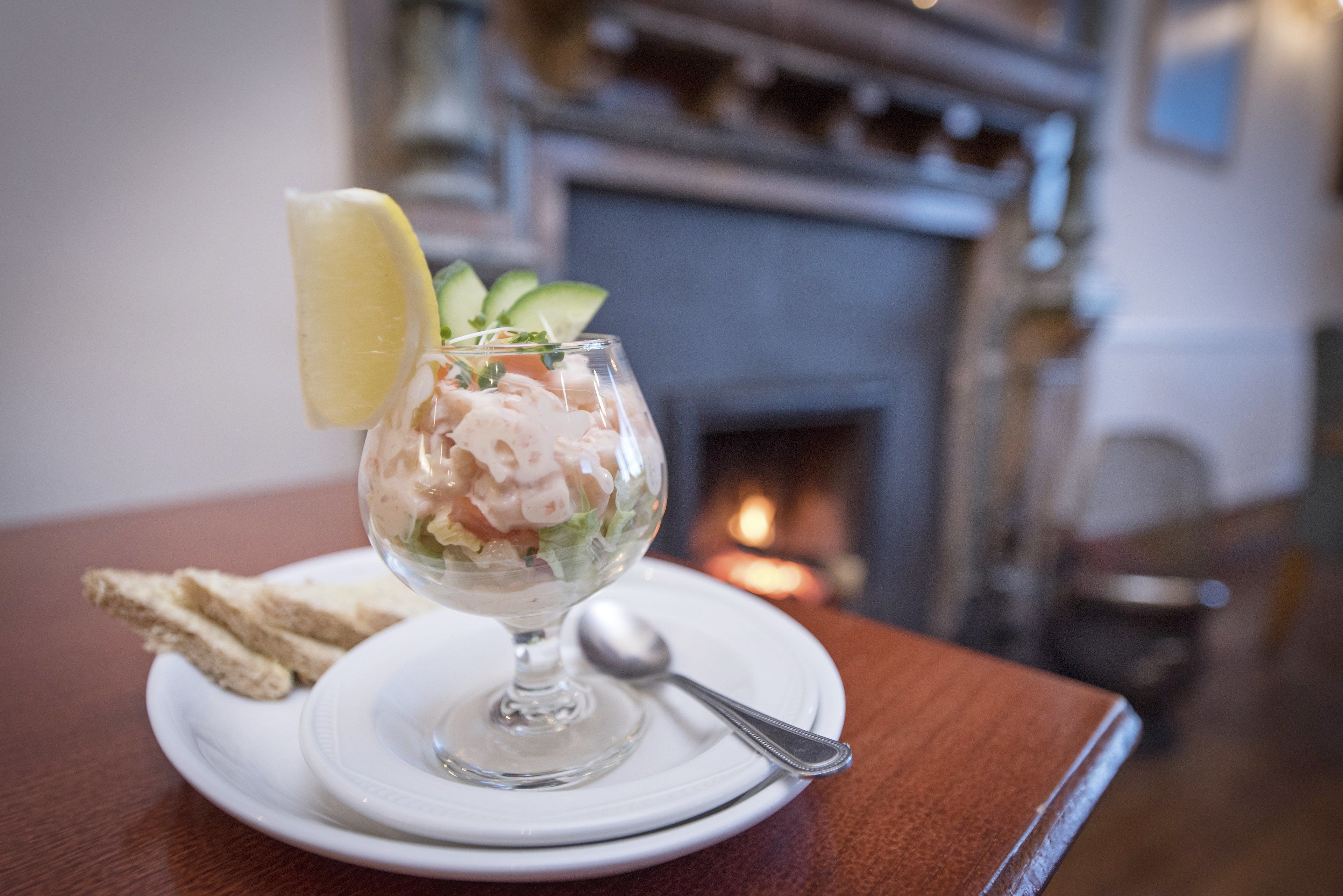 Een dessert in een glazen schaal op een wit bord met een lepel, vergezeld van sneetjes toast, geplaatst op een houten tafel met een open haard op de achtergrond.