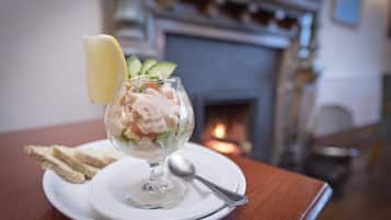 Een dessert in een glazen schaal op een wit bord met een lepel, vergezeld van sneetjes toast, geplaatst op een houten tafel met een open haard op de achtergrond.