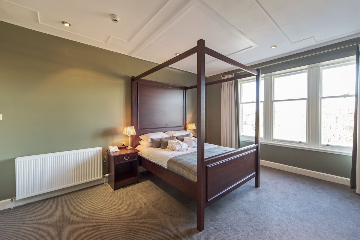 A four-poster bed in a room with a radiator, a nightstand with lamps, and a window with curtains.