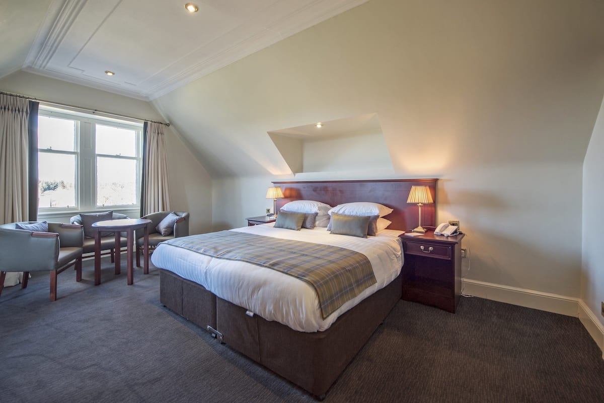 A hotel room with a large bed, a desk with a chair, a nightstand with a lamp, and a window with curtains.