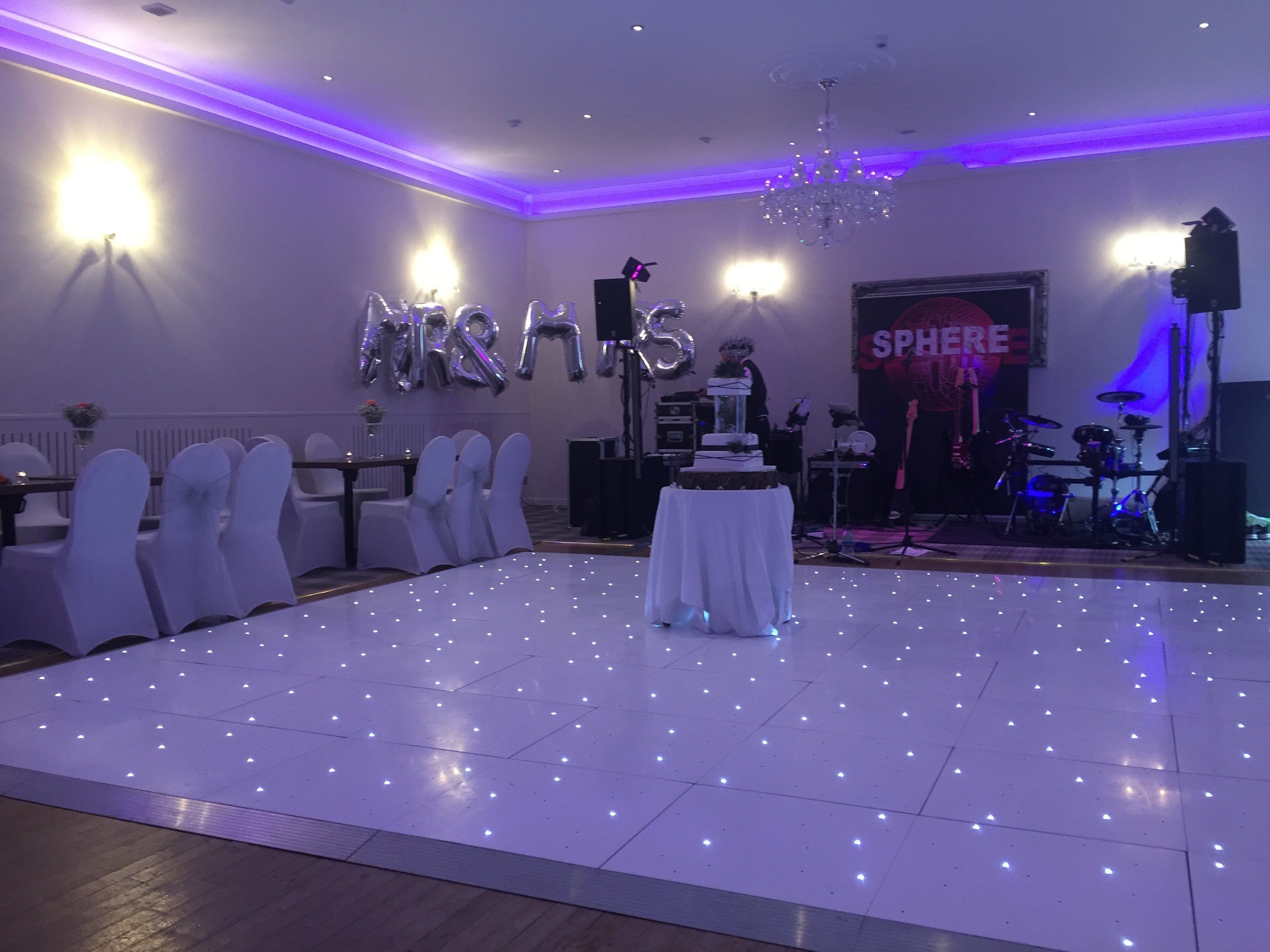 A wedding reception area with a dance floor, white chairs, a stage with musical instruments, and a 'MRS' balloon decoration.
