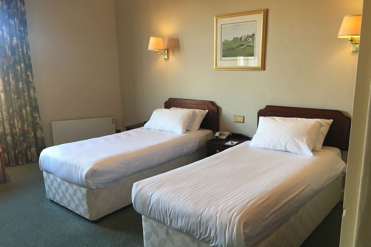 A hotel room with two single beds, a bedside table with a lamp, a painting on the wall, and a window with curtains.
