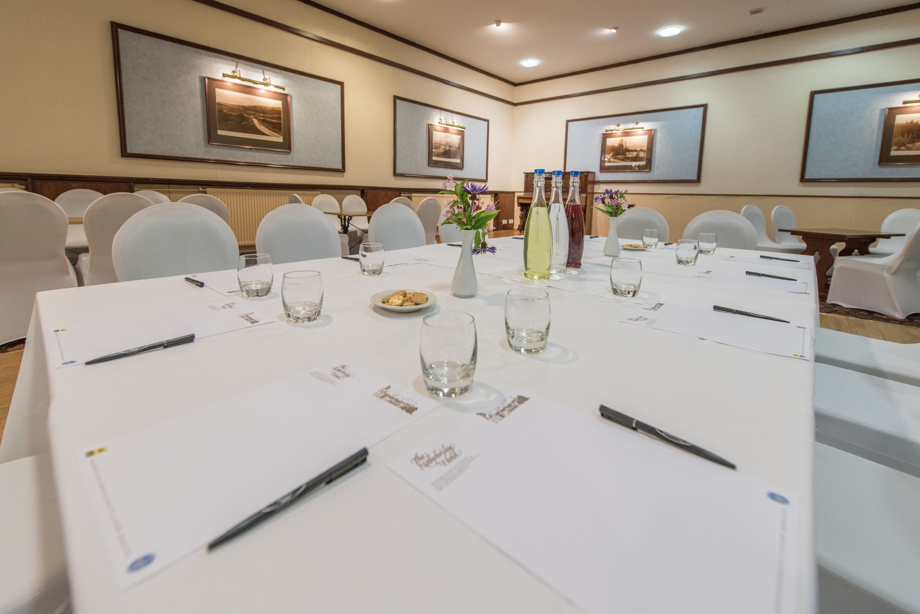 A conference room with white tables, chairs, and a white tablecloth. There are vases with flowers, water glasses, and pens on the tables.