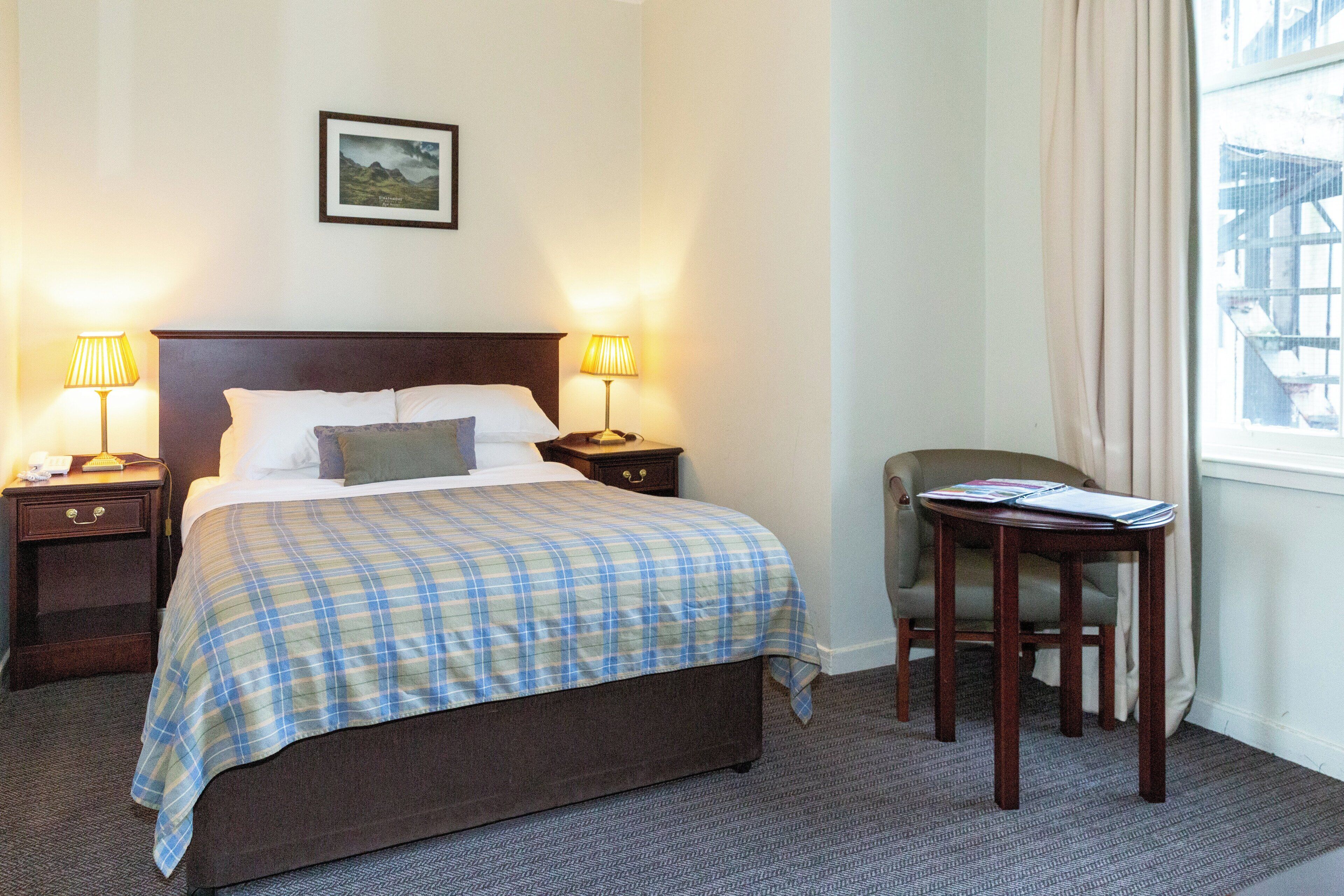 A hotel room with a large bed, two bedside tables with lamps, a chair, a small table with a magazine, and a window with curtains.