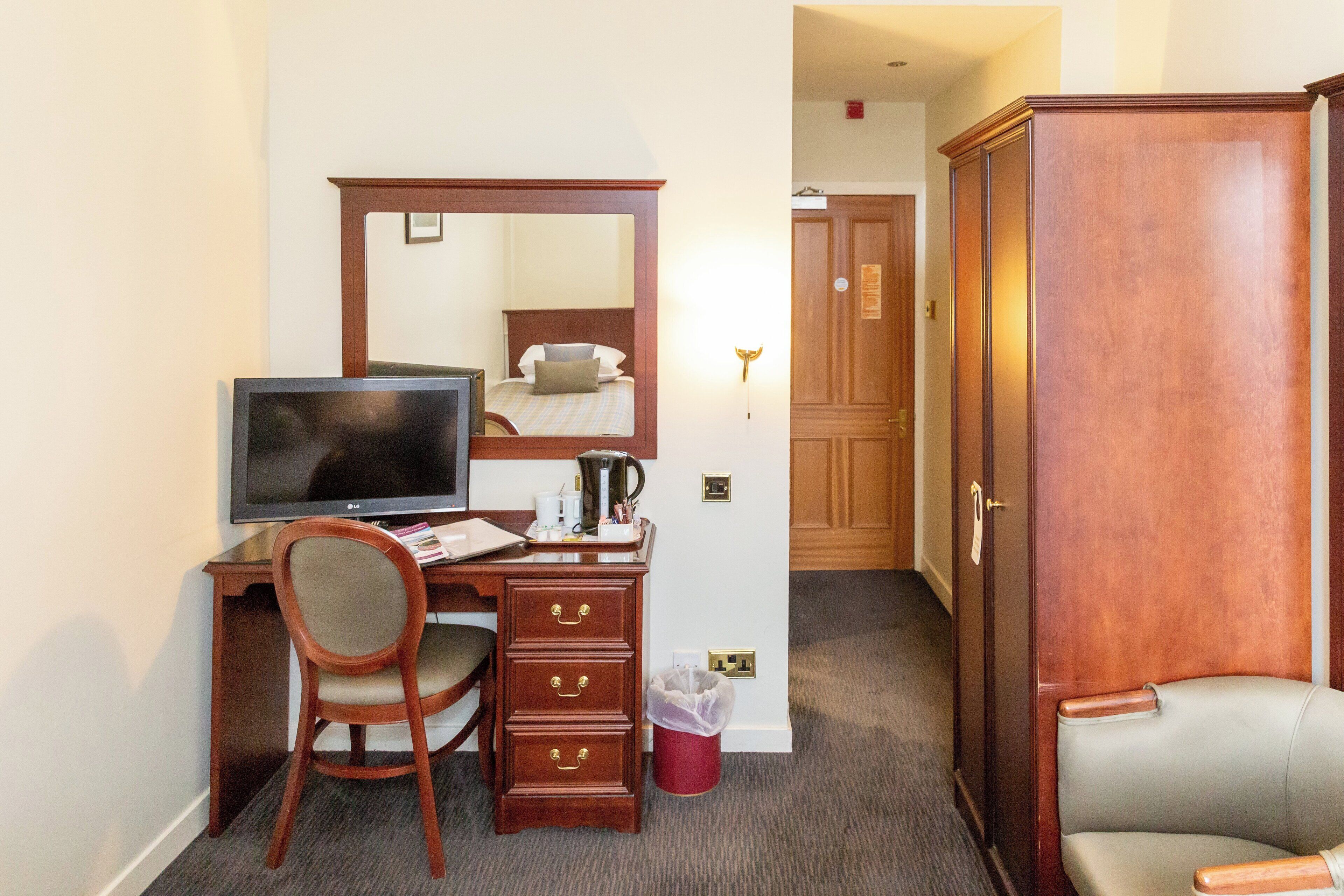 A hotel room with a wooden desk, a television, a chair, a wardrobe, and a trash bin.