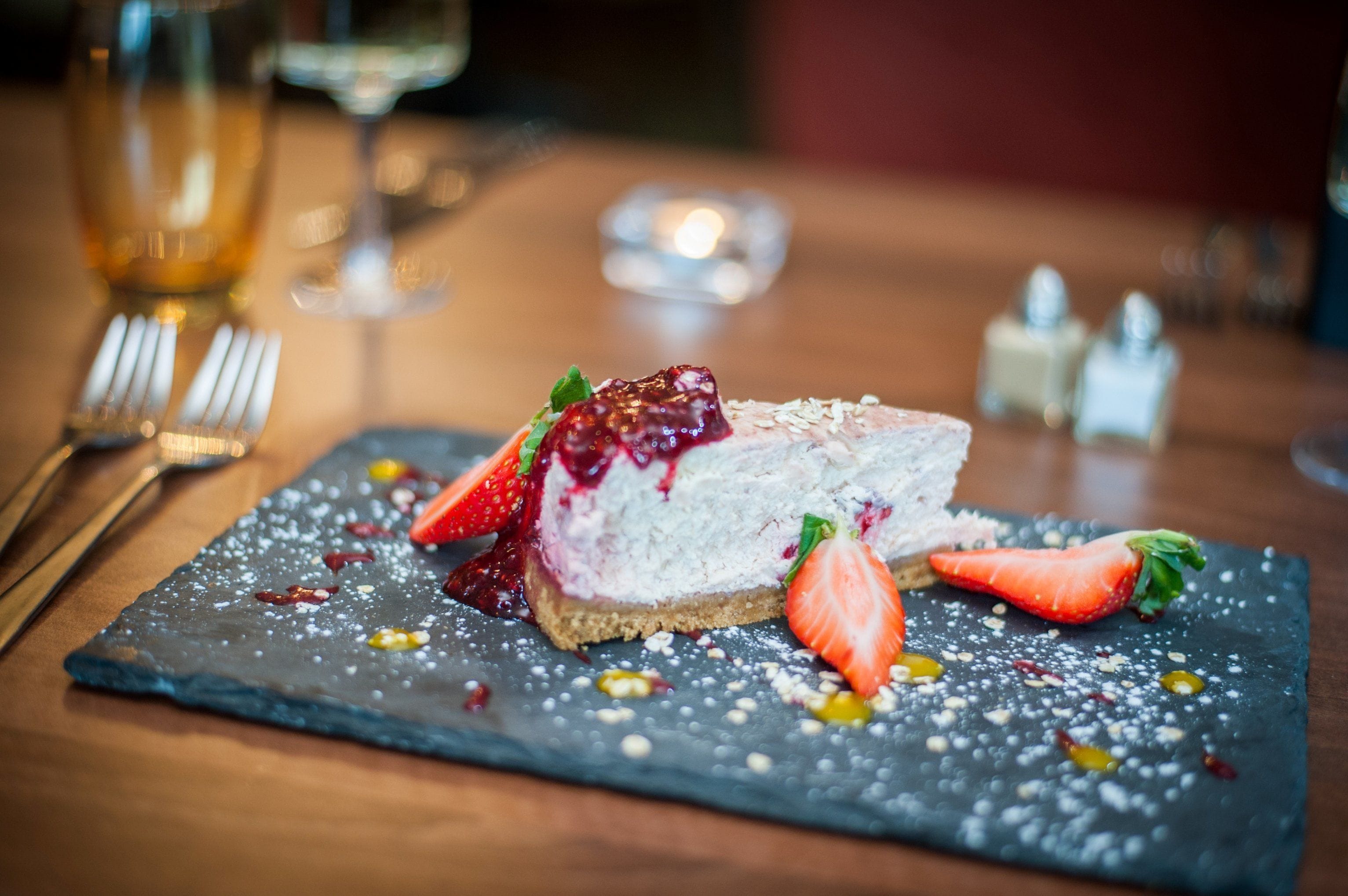 A dessert on a slate plate with strawberries and a berry compote, accompanied by a glass of wine and a candle in the background.