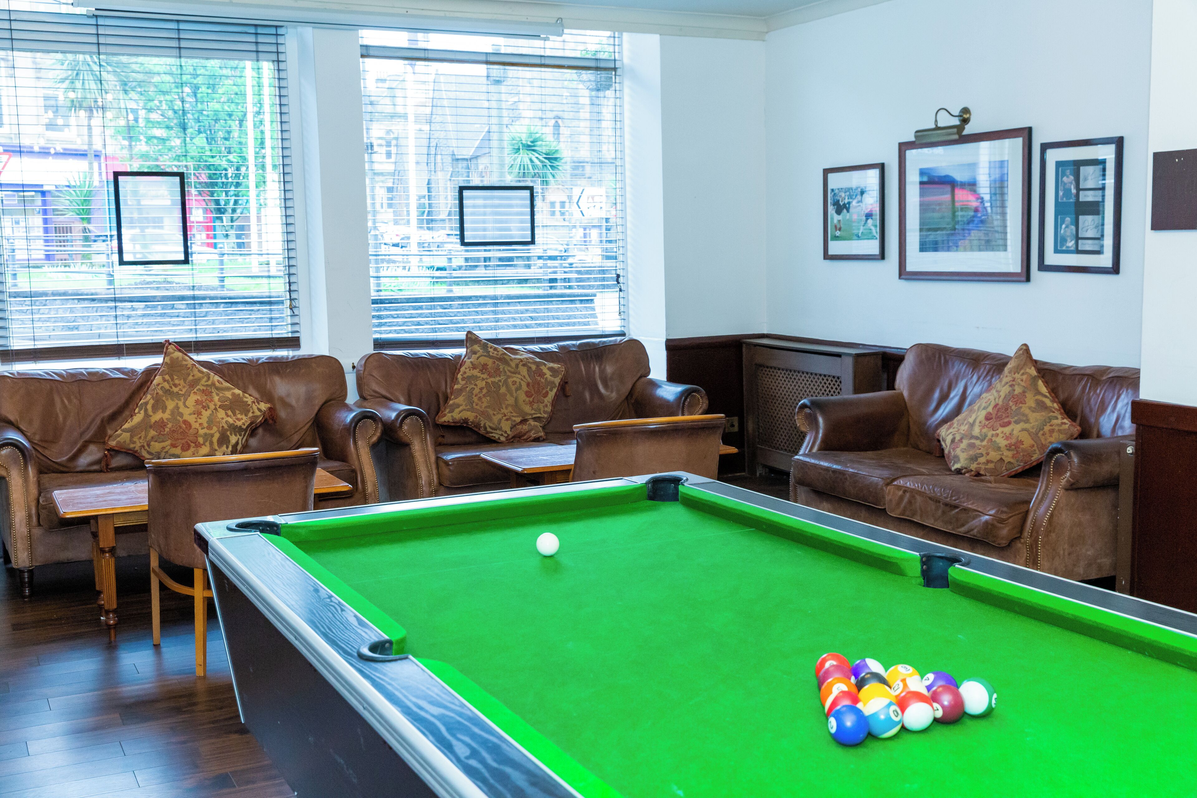 Une table de billard dans une pièce où se trouvent des canapés et des fauteuils en cuir, une queue de billard et des tableaux encadrés accrochés au mur.