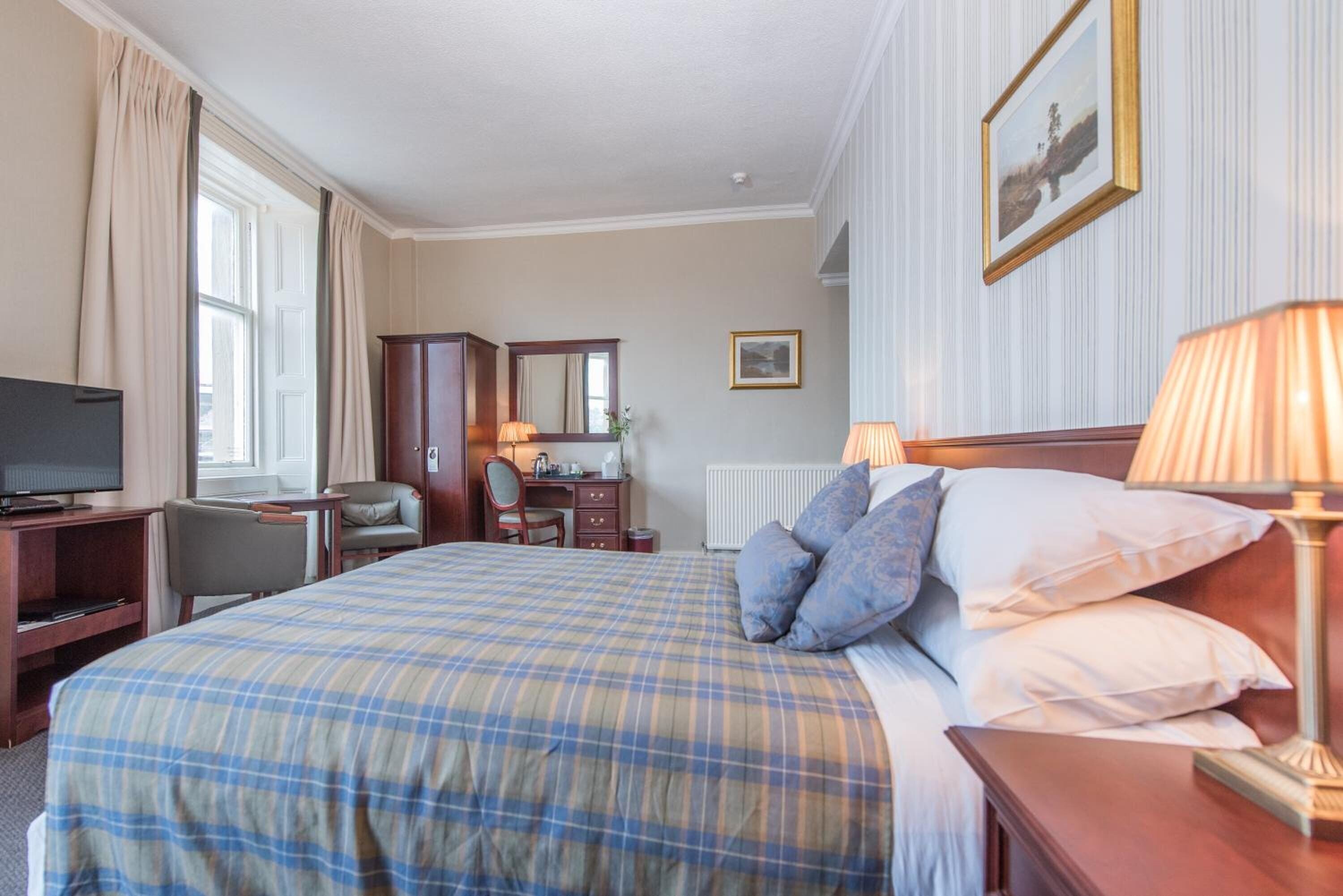 A hotel room with a large bed, a desk with a computer, a chair, a television, and a window with curtains.