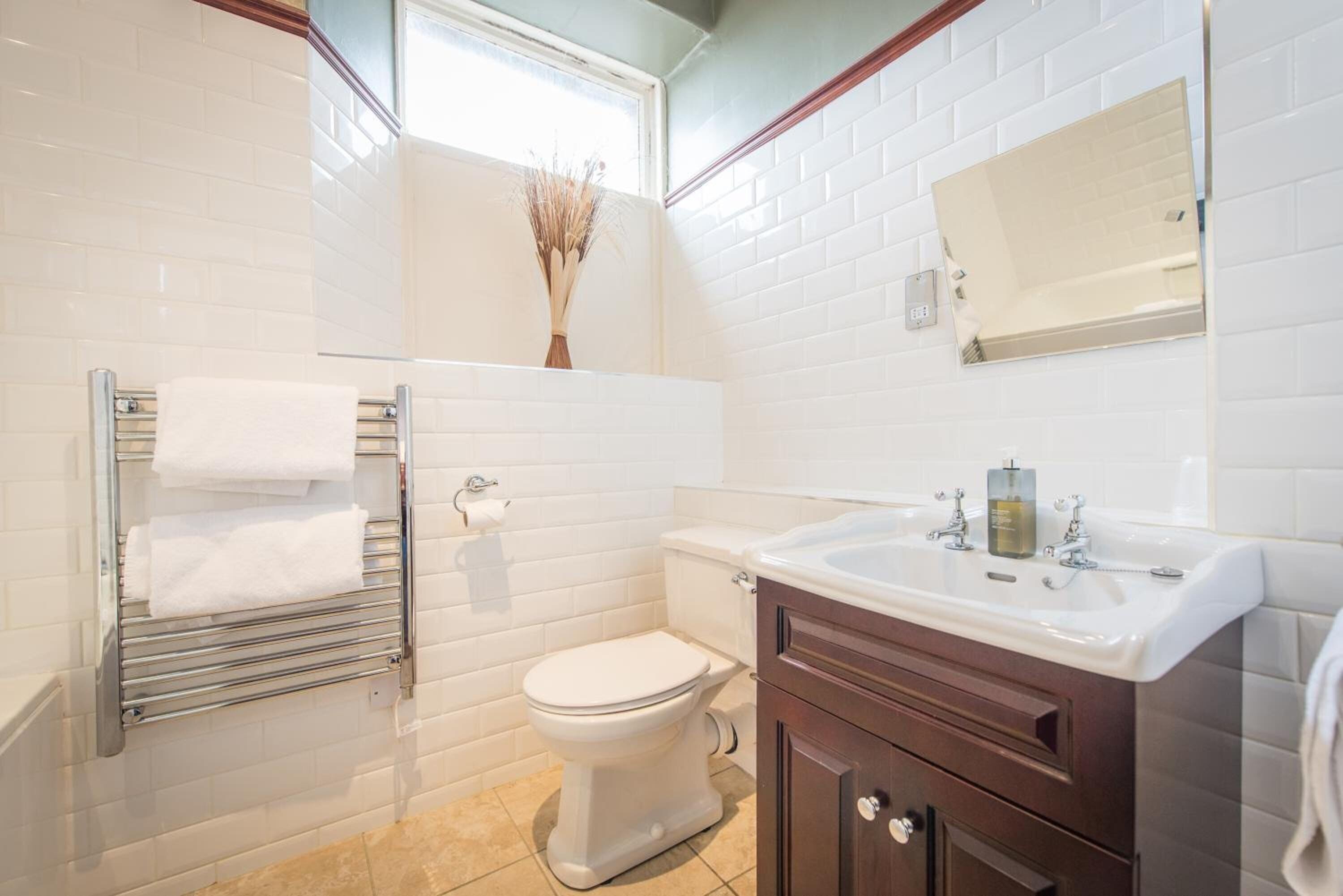 A bathroom with white subway tiles, a white toilet, a glass shower enclosure, a towel rack with white towels, a wooden vanity with a mirror, and a bottle of hand soap on the sink.
