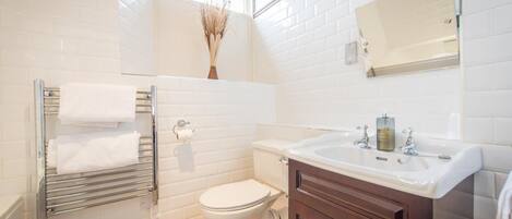 A bathroom with white subway tiles, a white toilet, a glass shower enclosure, a towel rack with white towels, a wooden vanity with a mirror, and a bottle of hand soap on the sink.