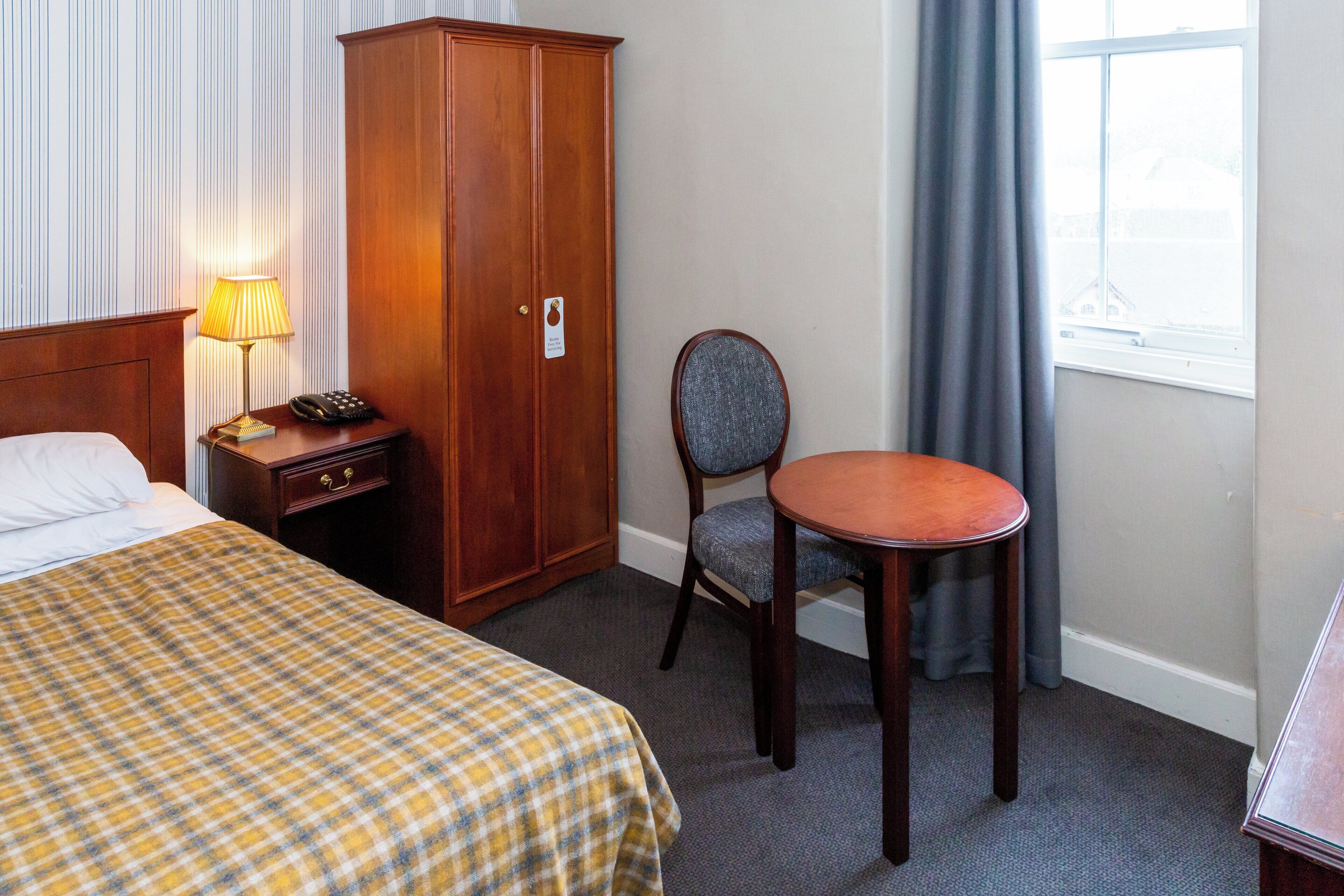 A hotel room with a bed, a desk with a lamp, a chair, a wardrobe, and a window with curtains.