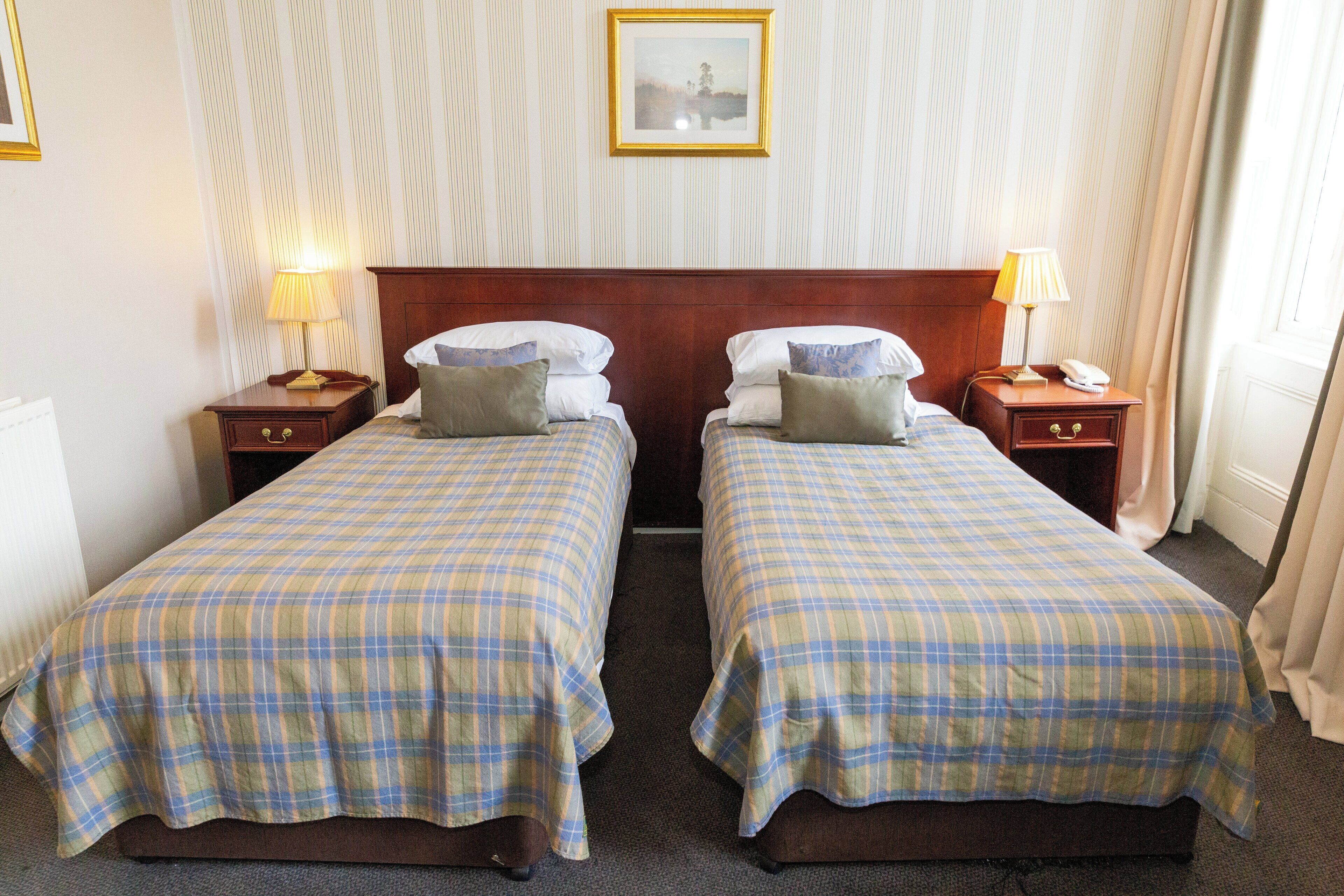 A hotel room with two beds, each with a plaid bedspread, wooden headboards, bedside tables with lamps, and a framed picture on the wall.