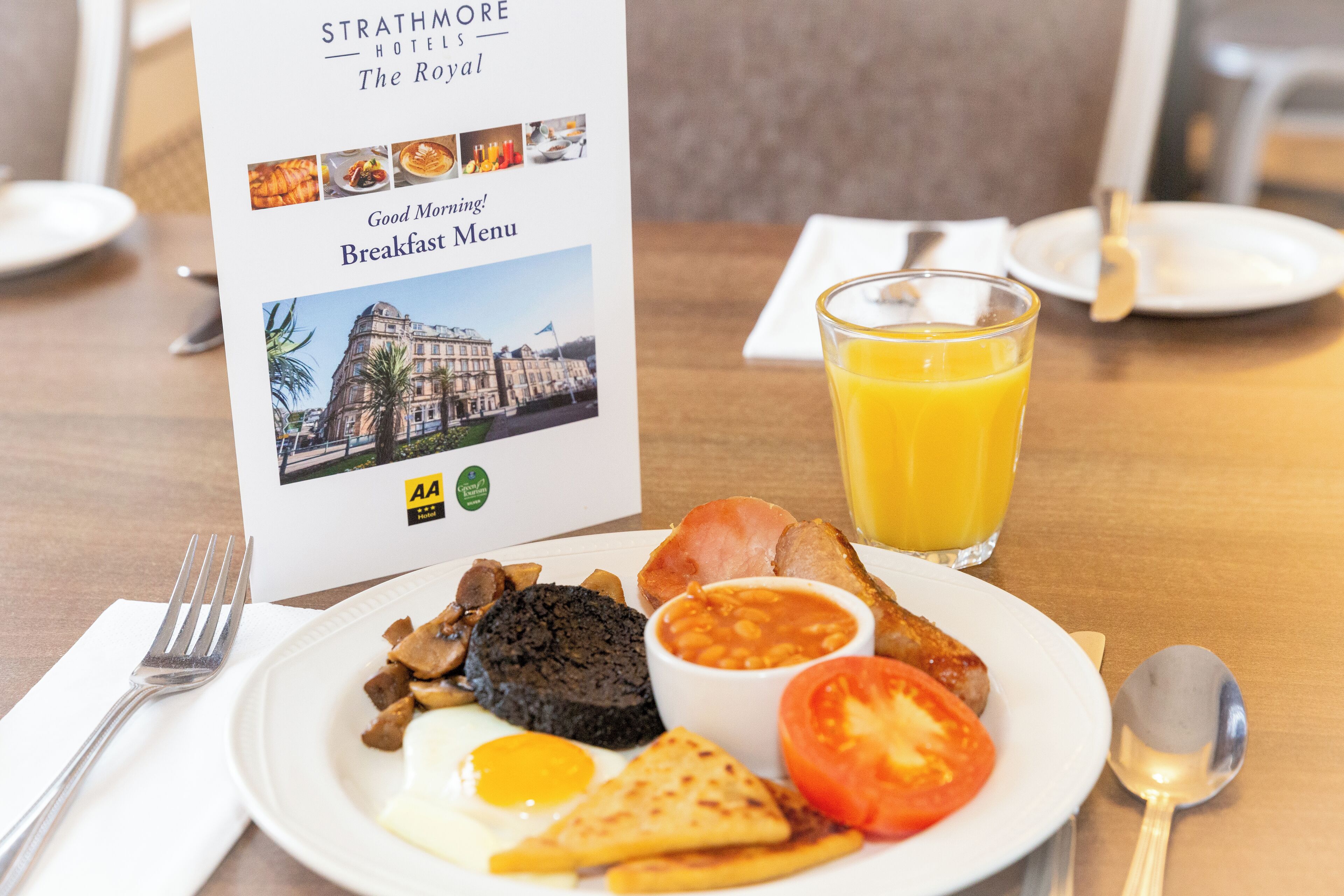 A breakfast plate with eggs, beans, tomatoes, and toast, accompanied by a glass of orange juice and a menu from Strathmore Hotels.