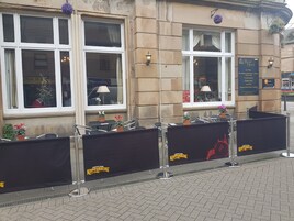 Exterior de un hotel con amplios ventanales, zona de estar al aire libre y una barrera negra con la marca 'Kopparberg'.