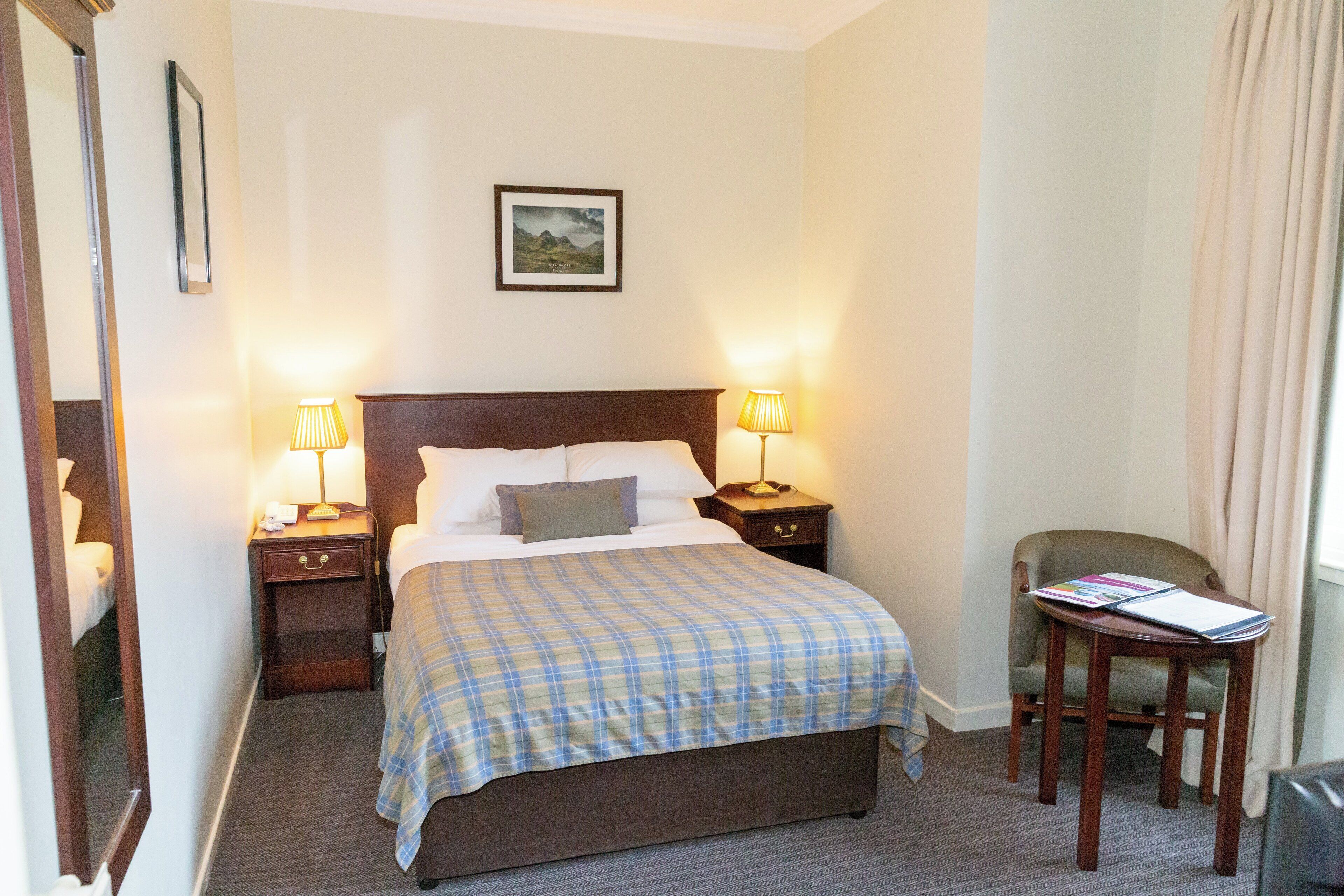 Ein Hotelzimmer mit einem großen Bett, zwei Nachttischen mit Lampen, einem Sessel und einem kleinen Tisch mit einer Zeitschrift.
