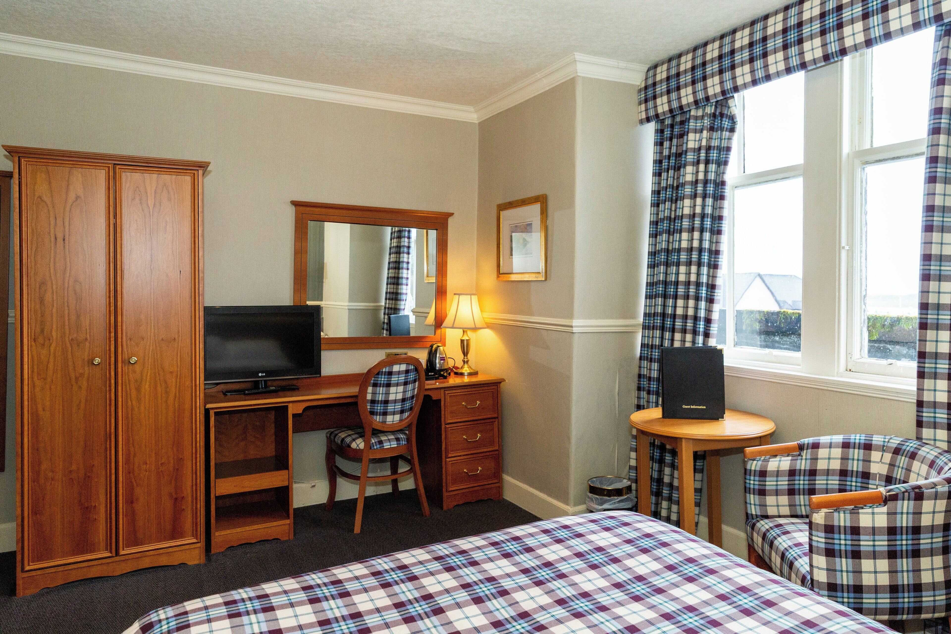 A hotel room with a bed, a desk with a computer, a chair, a wardrobe, a mirror, and a window with curtains.