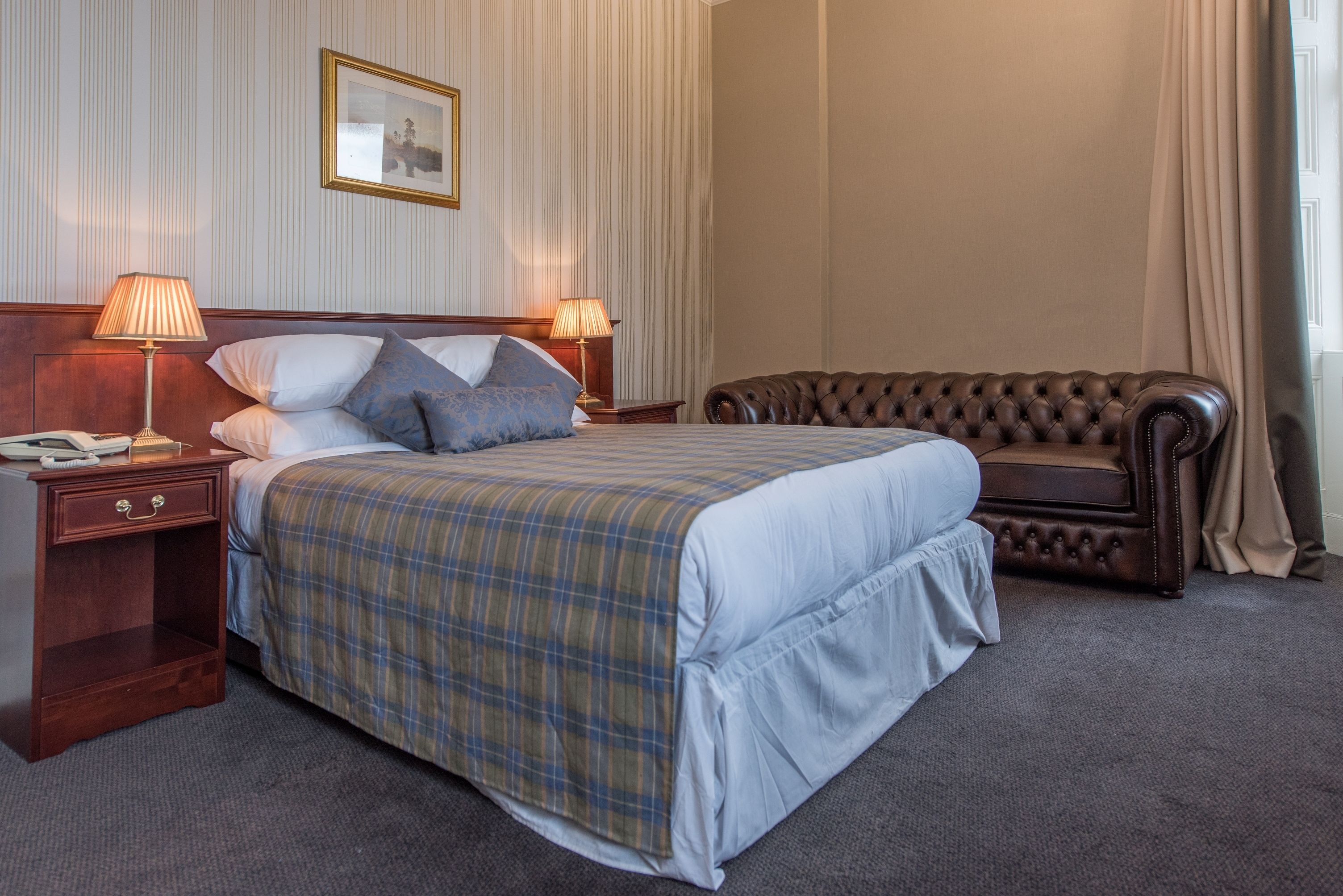 A hotel room with a large bed, two bedside lamps, a wooden nightstand, a leather sofa, and a framed picture on the wall.