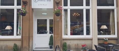 A building with a sign for 'Strathmore Hotels Lounge Bar @ The Royal'. There are steps leading to the entrance, a small potted plant on the steps, and a chair and table outside.