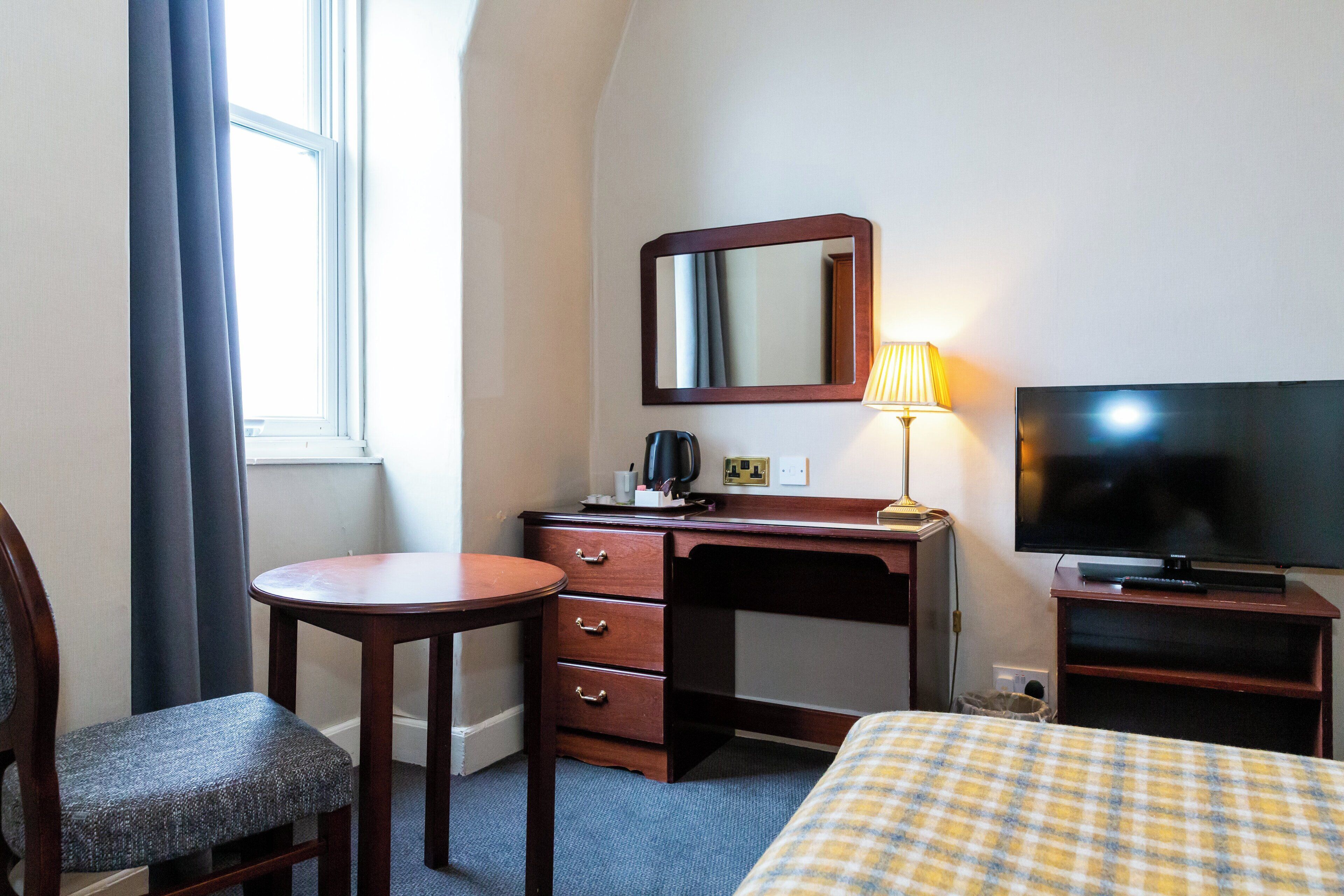 Quarto de hotel com uma cama, uma escrivaninha com cadeira, televisão e janela com cortinas.
