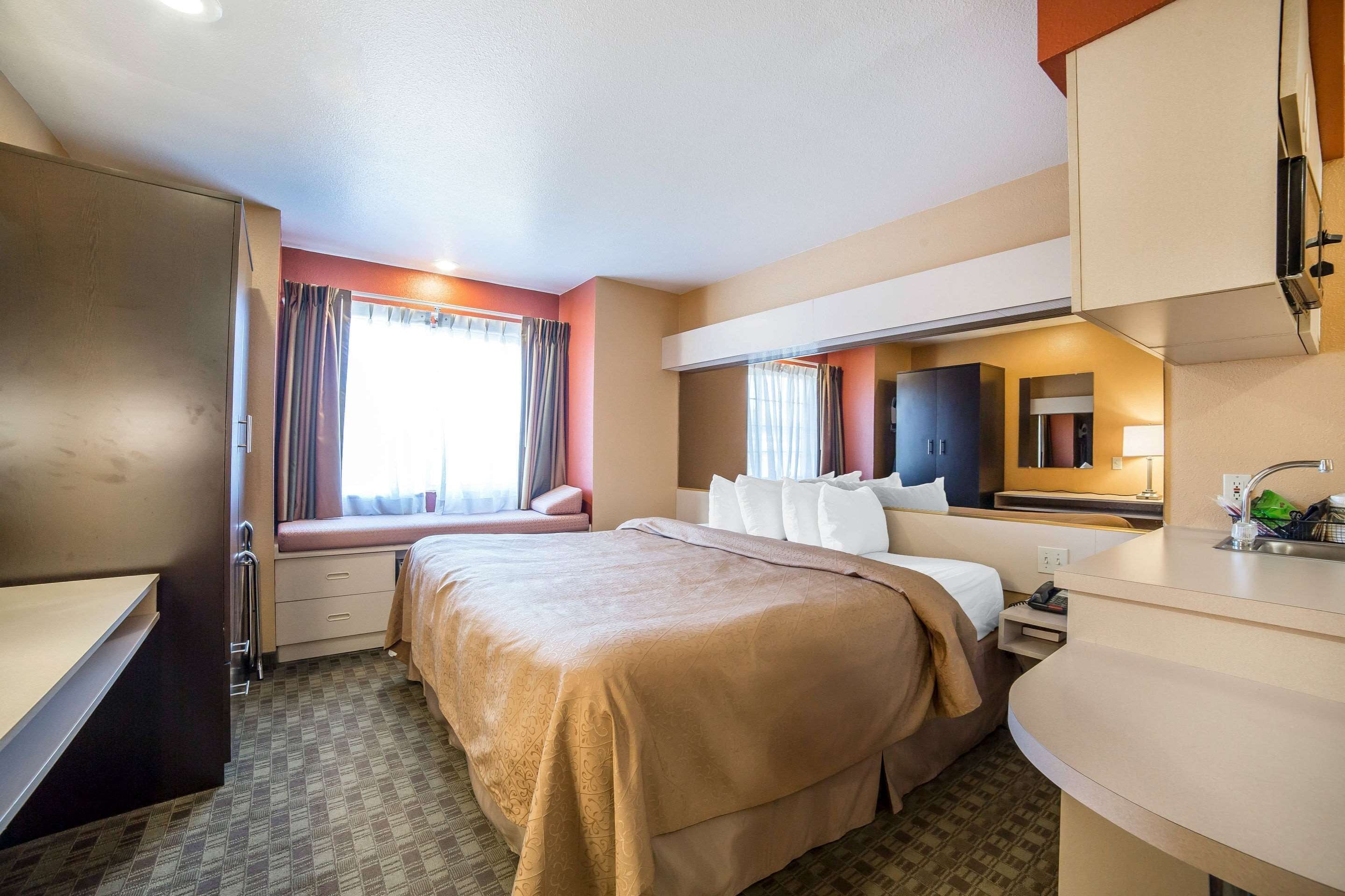 A hotel room with a bed, a desk, a dresser, a window with curtains, and a bathroom area.