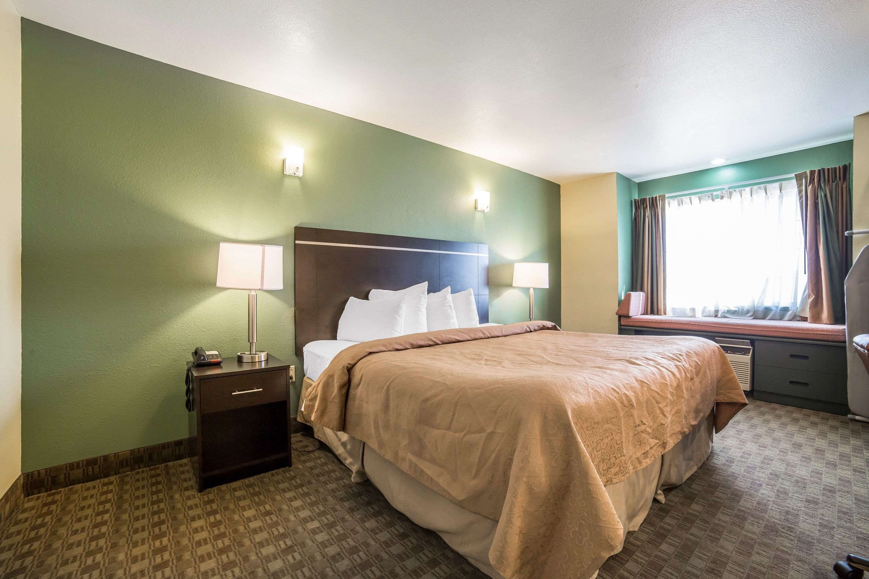 A hotel room with a bed, bedside lamps, a nightstand, a window with curtains, and a view of the outside.