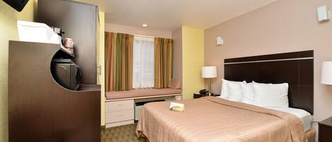 A hotel room with a bed, a desk, a television, a window with curtains, and a nightstand with a lamp.