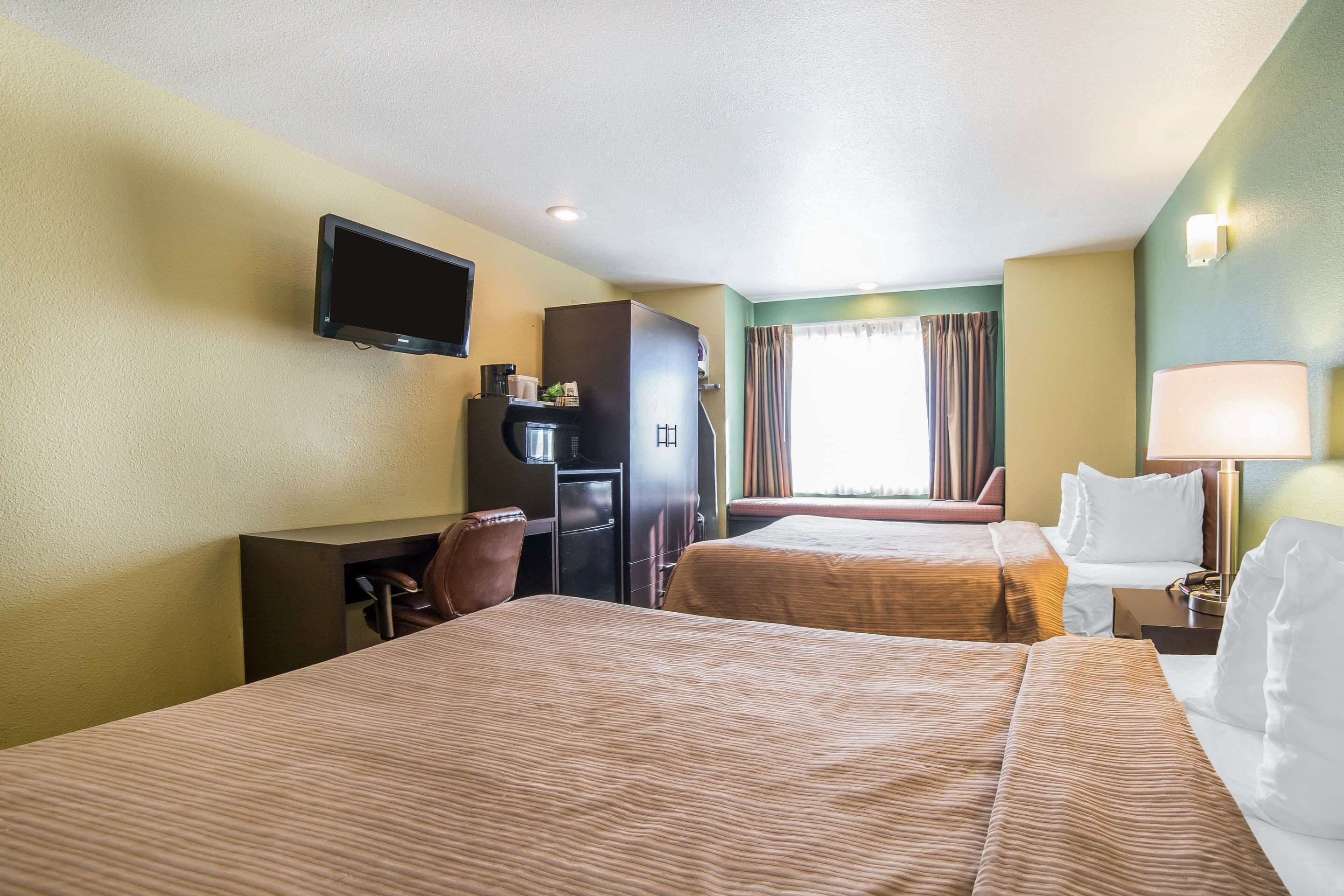 A hotel room with two beds, a desk, a television, a refrigerator, and a window with curtains.