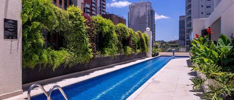 Ein Schwimmbecken mit einer Metallleiter, umgeben von einer gefliesten Terrasse und üppigem Grün.