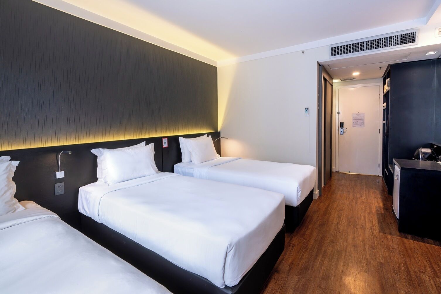 Quarto de hotel com três camas de solteiro, cabeceira escura, piso de madeira e uma pequena área de cozinha.