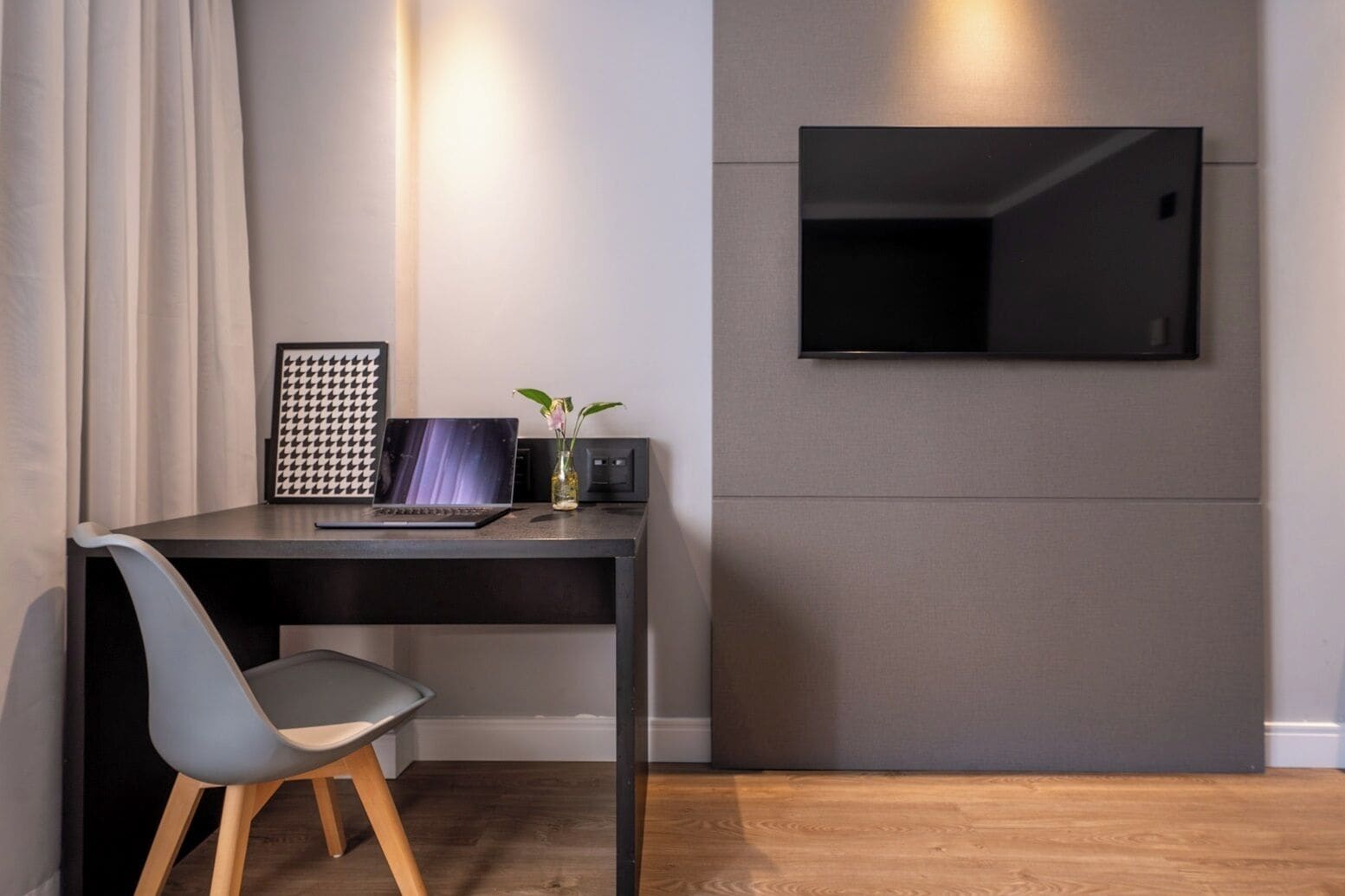 Une chambre moderne avec un bureau sombre, un ordinateur portable, un tableau encadré et un petit vase avec des fleurs. Un téléviseur à écran plat est fixé au mur.