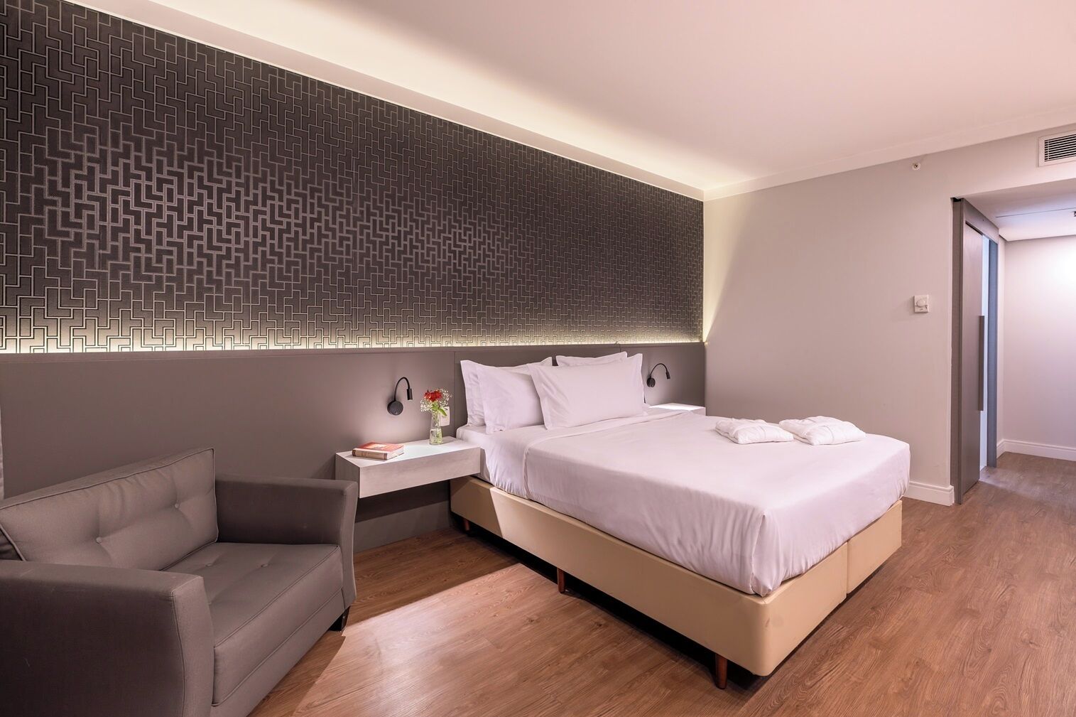 A modern hotel room with a large bed, a bedside table with a lamp, a chair, and a patterned wall.