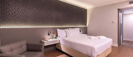 A modern hotel room with a large bed, a bedside table with a lamp, a chair, and a patterned wall.