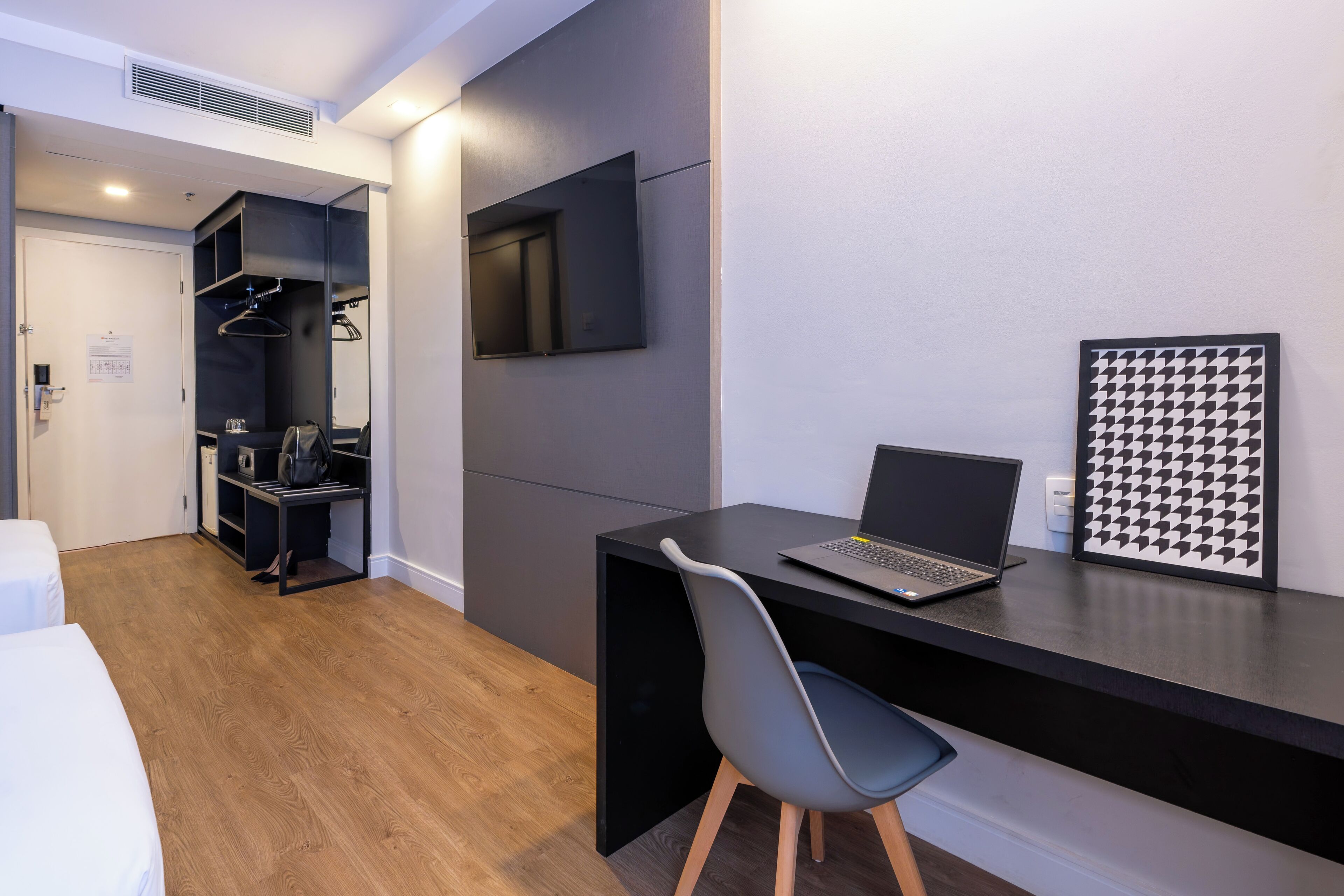 A modern room with a black desk, a laptop, a grey chair, a black and white patterned artwork, and a wardrobe with a mirror.