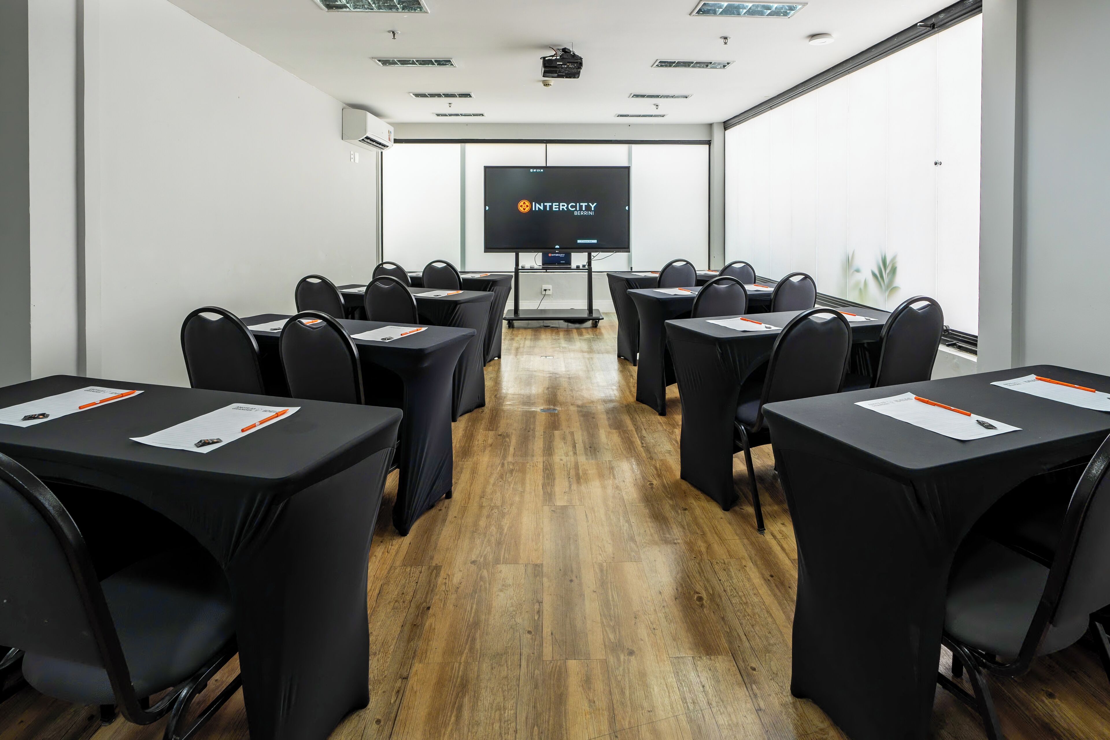 A conference room with a large screen displaying "INTERCITY" and "SEMINAR". Tables are arranged in a U-shape with black chairs.