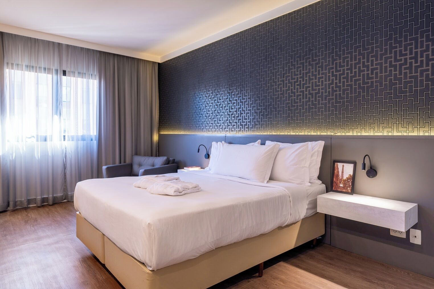 Habitació d'hotel moderna amb un llit gran, una taula de nit, una cadira i una paret amb un disseny geomètric.