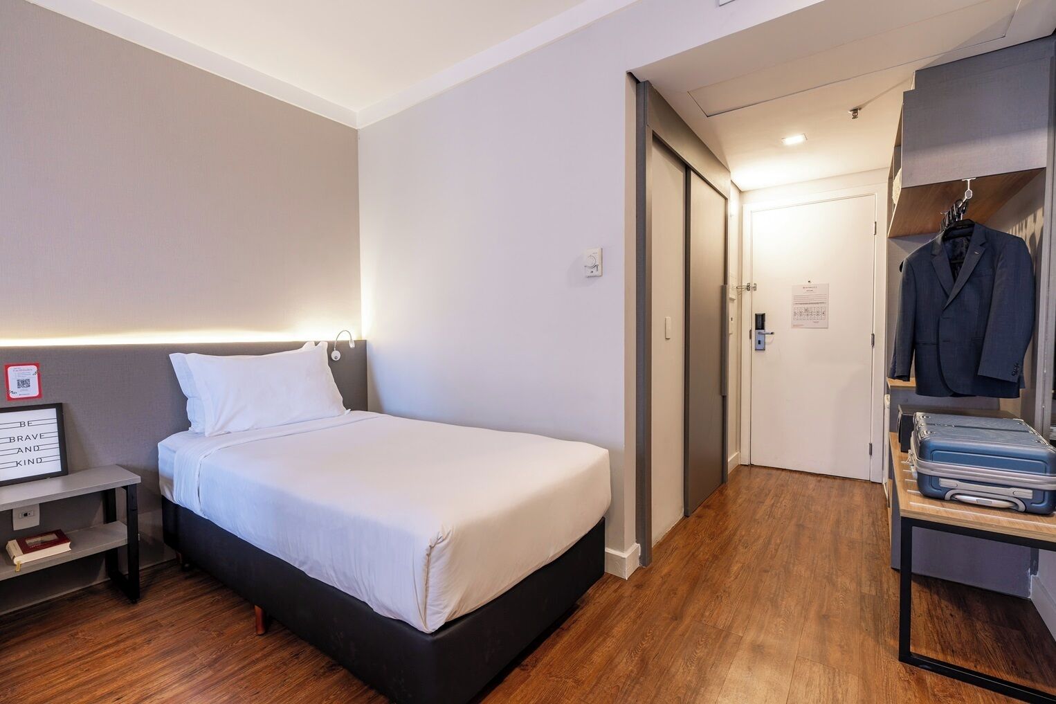 Une chambre d’hôtel avec un lit, une table de chevet et une armoire contenant des vêtements suspendus et une valise.